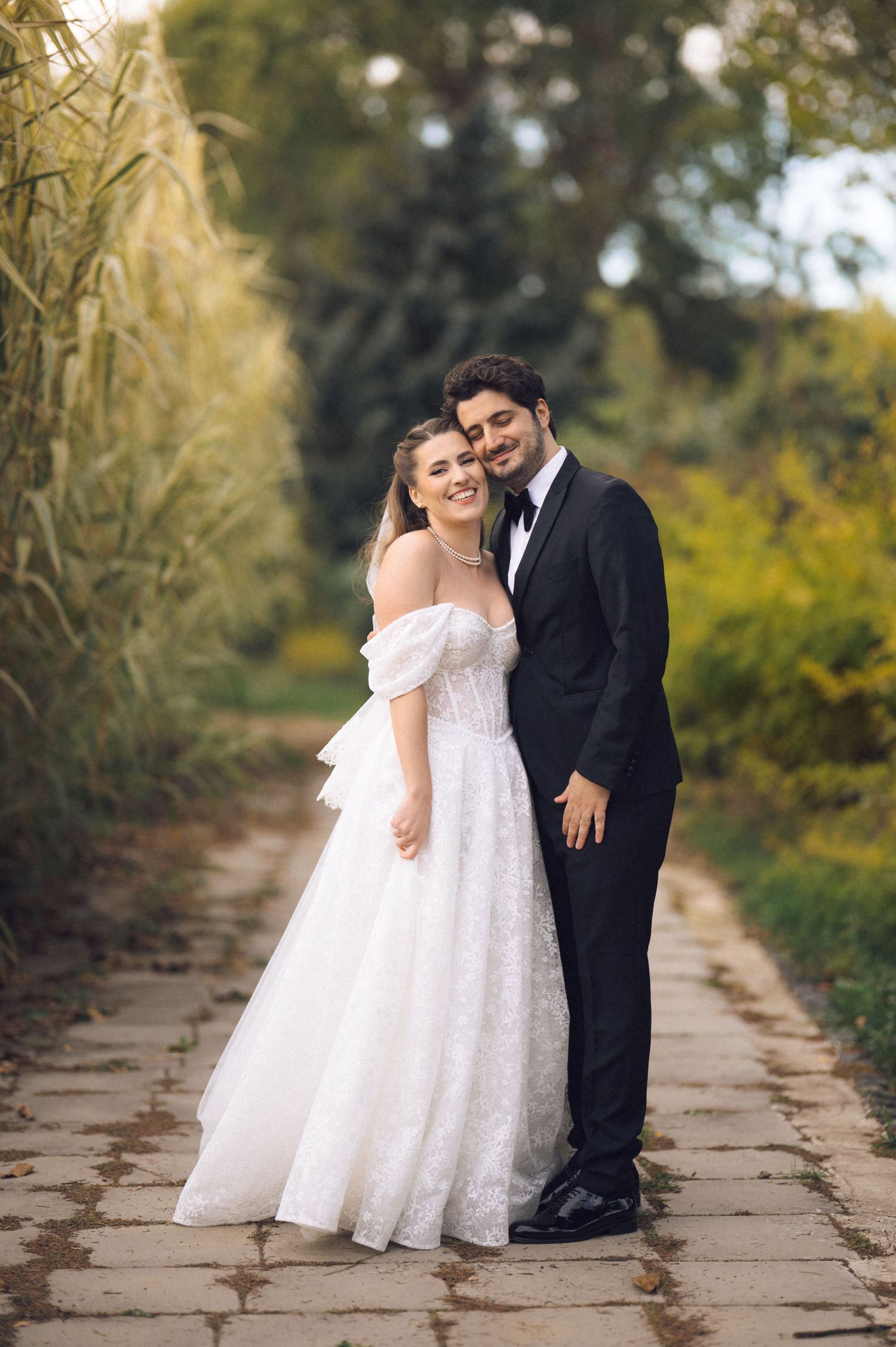 A couple in formal wedding attire shares a tender moment on a stone path lined with tall bamboo plants.
