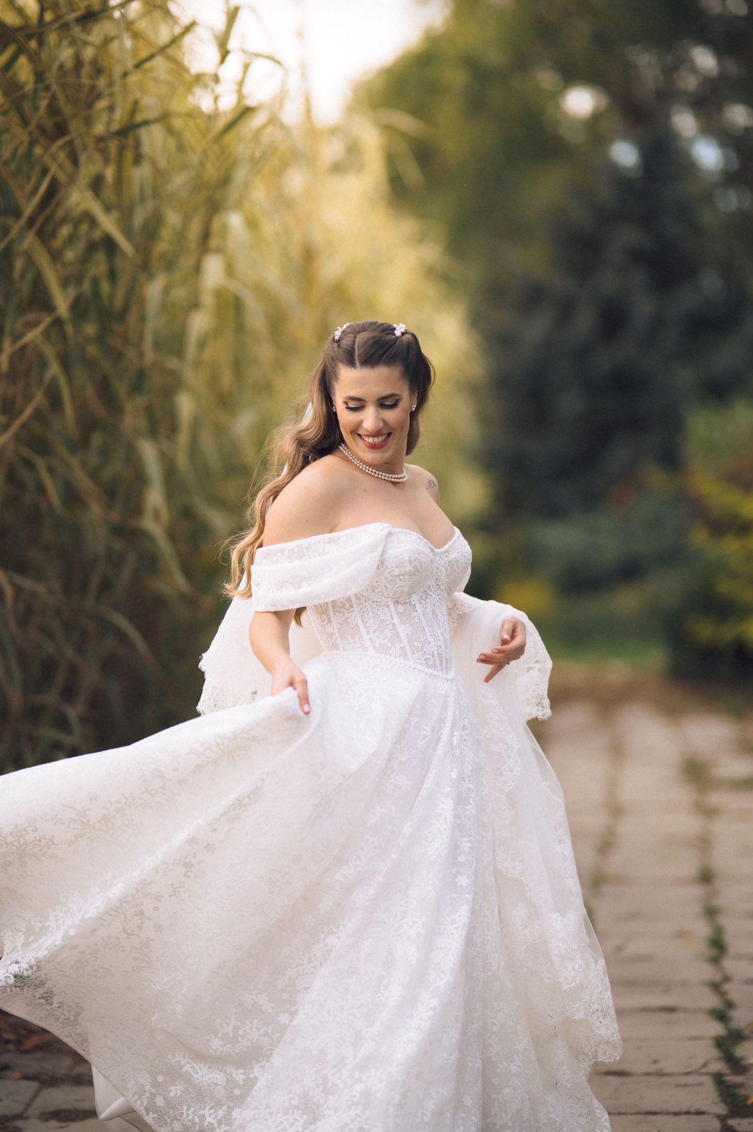 A bride in a white lace off-shoulder gown twirls joyfully on a stone path during a sunset photoshoot.
