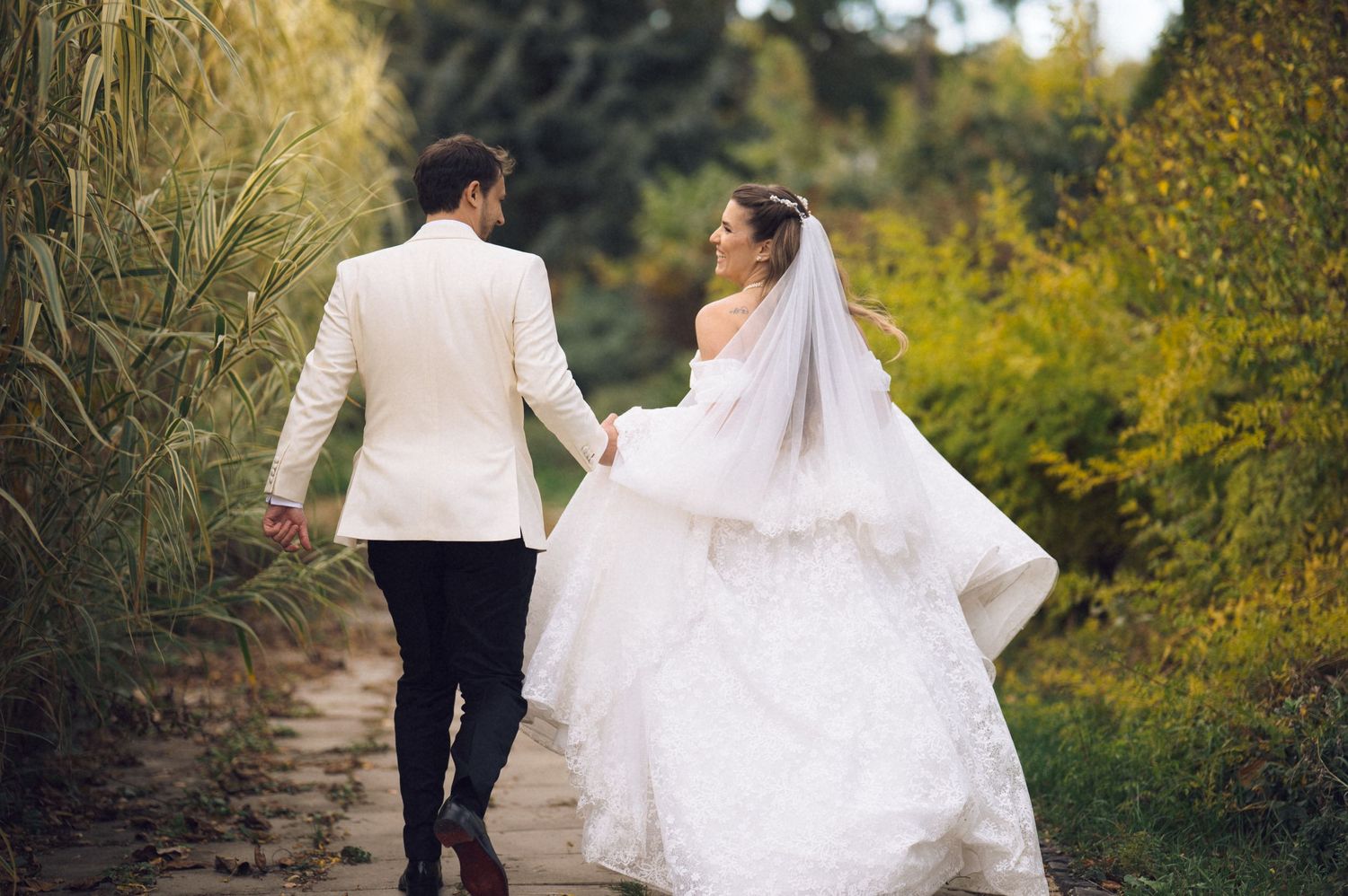 A bride and groom walk hand in hand through a garden path surrounded by autumn foliage.