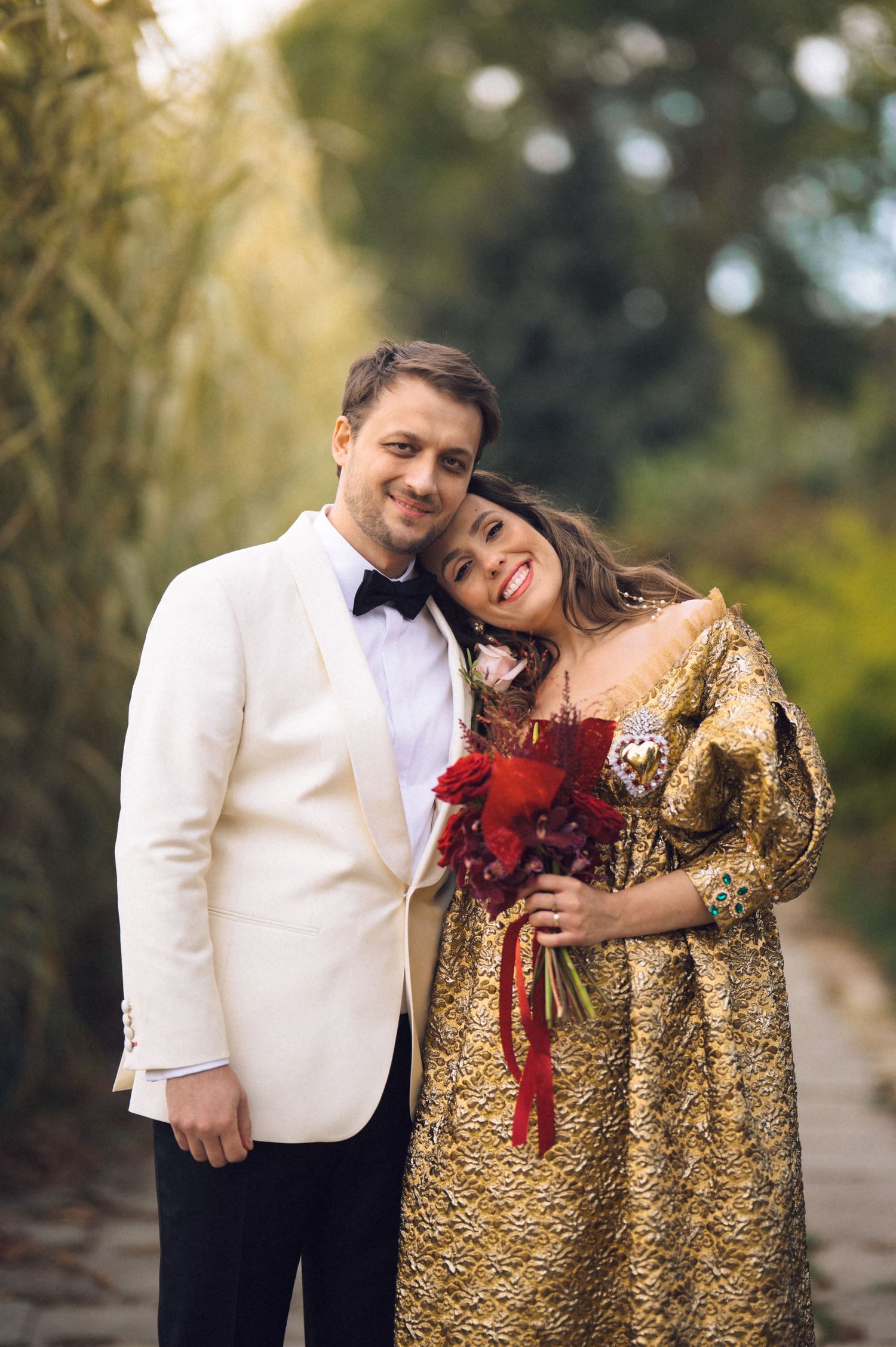 A couple poses outdoors in formal attire with a white tuxedo jacket and gold sequined dress for romantic photos.