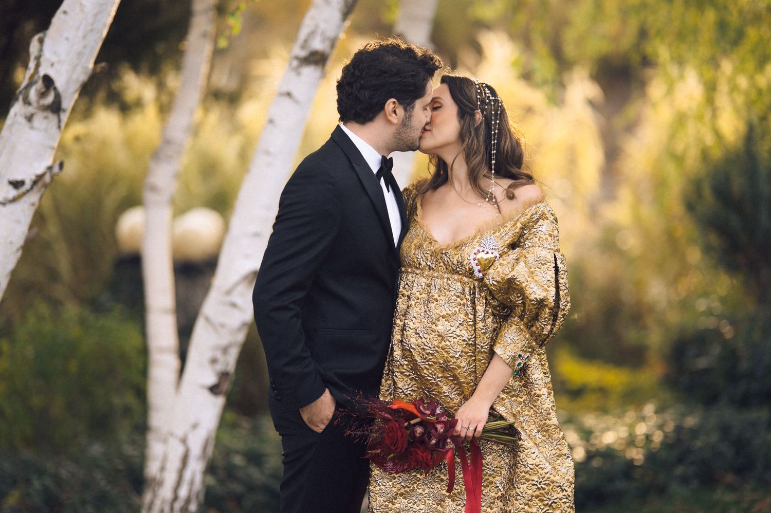 A couple in formal attire shares a romantic moment in a birch tree forest during golden hour.
