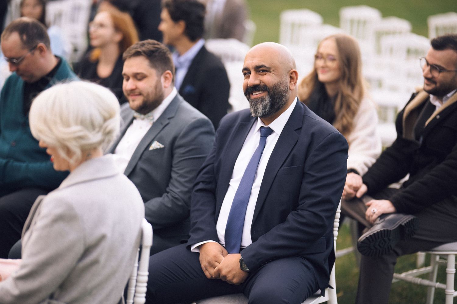 Wedding guests seated in white chairs at an outdoor ceremony share a friendly conversation before the event begins.