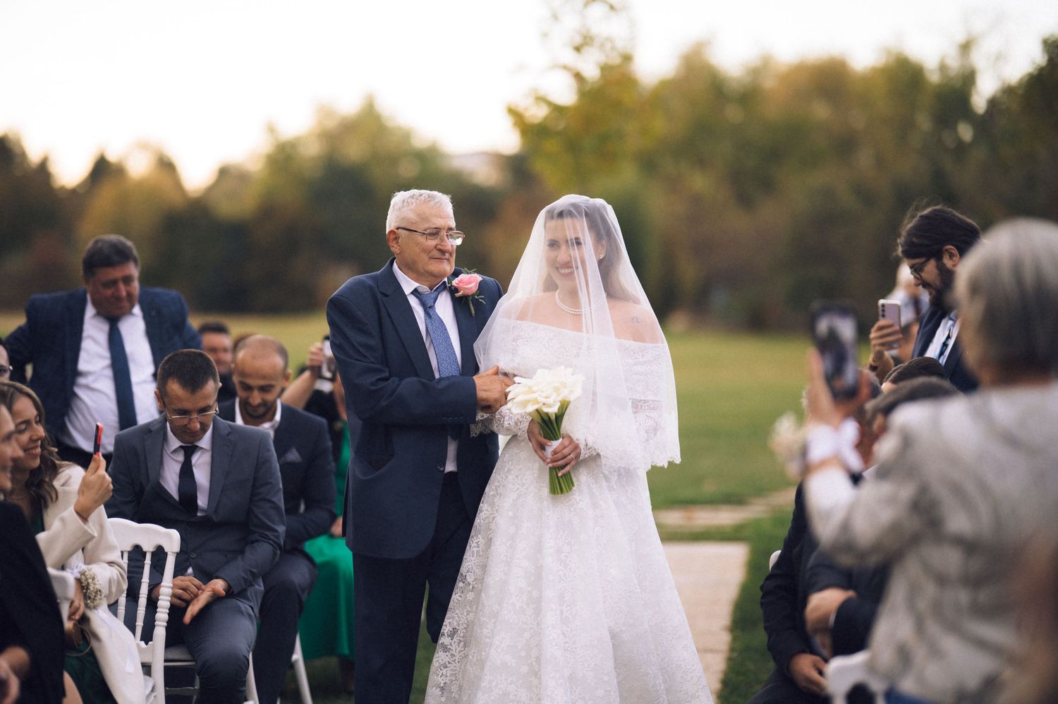 An outdoor wedding ceremony takes place as someone walks down the aisle in a white lace gown and veil at sunset.