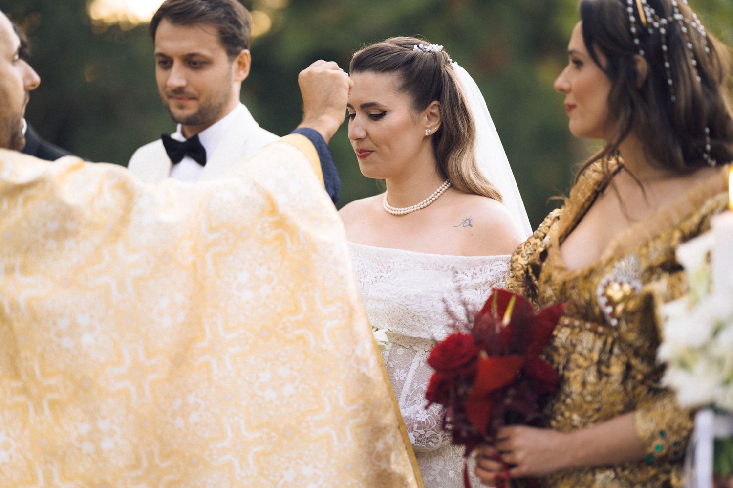 A wedding ceremony outdoors with people gathered in formal attire including white and gold dresses and a tuxedo.