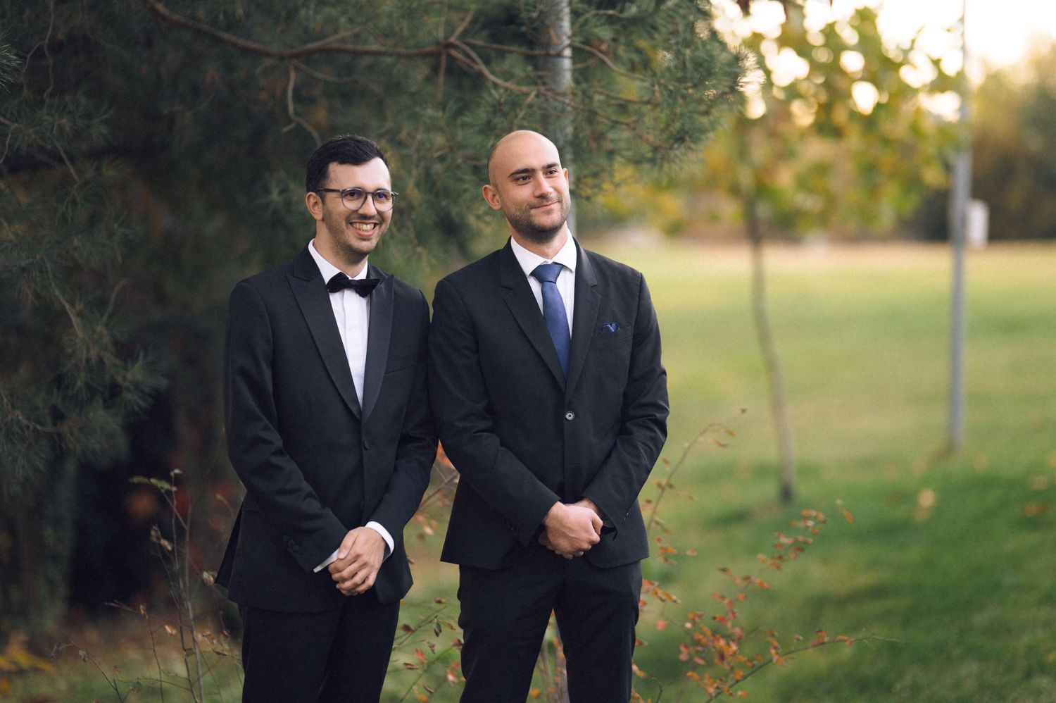Two well-dressed men in formal attire stand together outdoors during golden hour in a park setting.