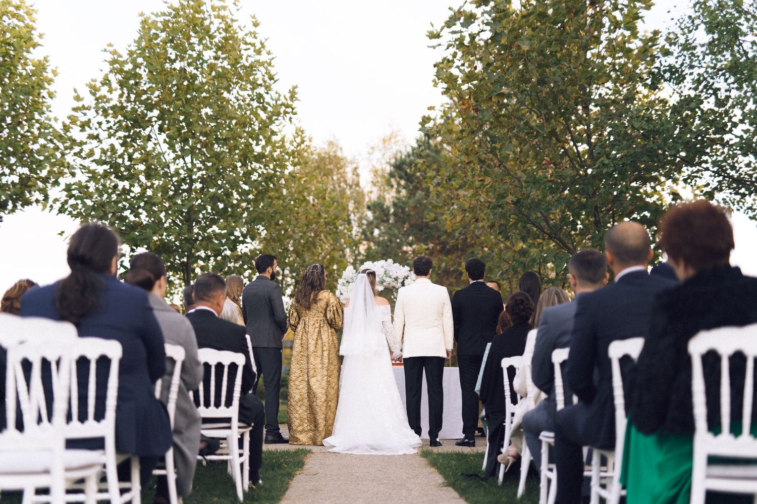 Wedding guests stand in a row outdoors during a ceremony with white chairs and autumn trees in the background.