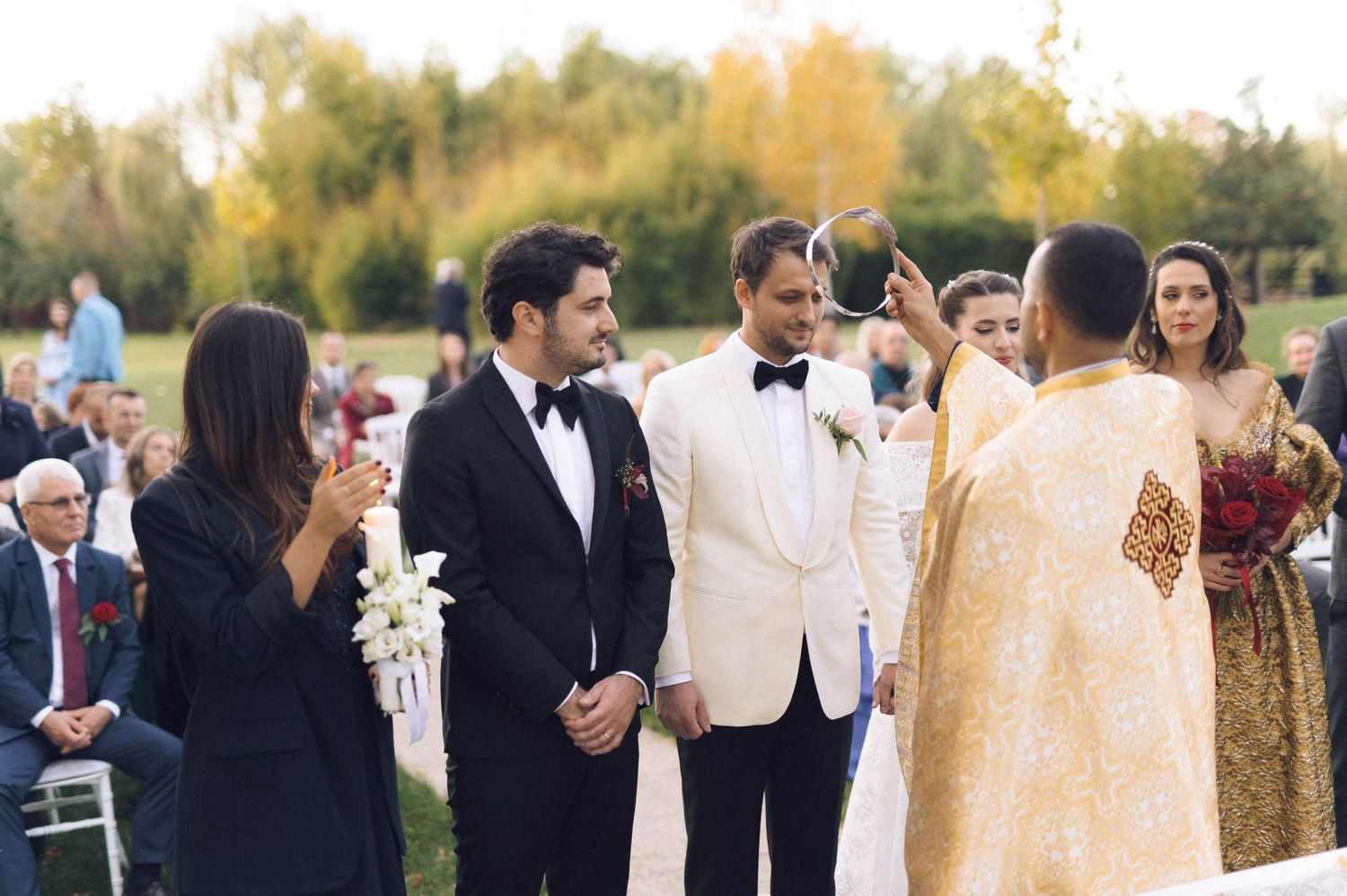 Outdoor wedding ceremony takes place in autumn with guests gathered in formal attire against golden foliage backdrop.