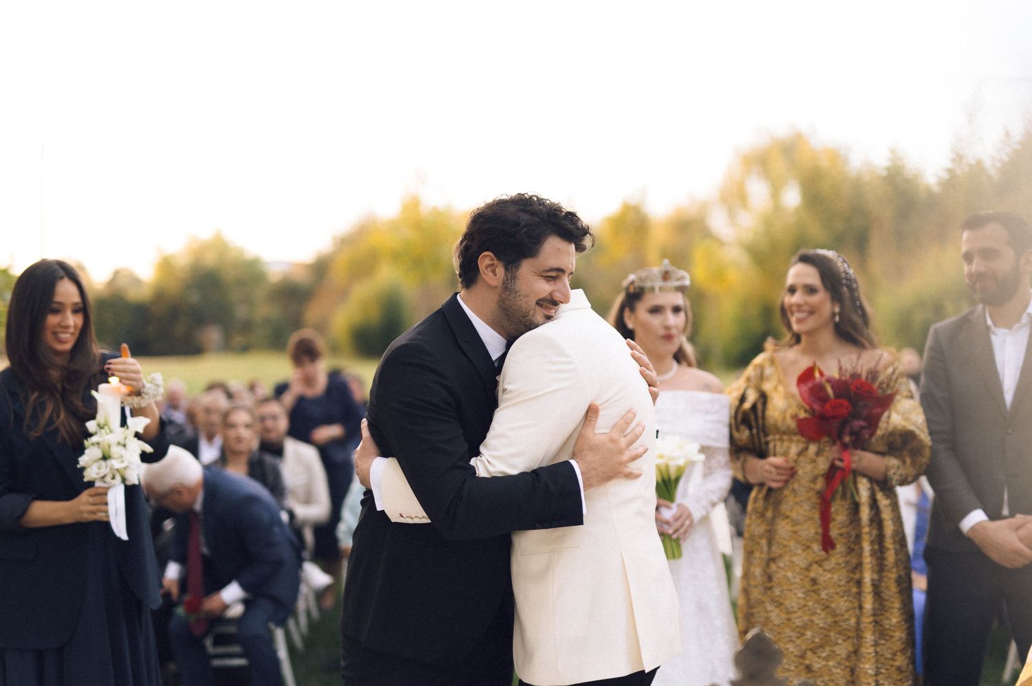 A couple shares their first dance at an outdoor wedding celebration surrounded by guests and autumn foliage.