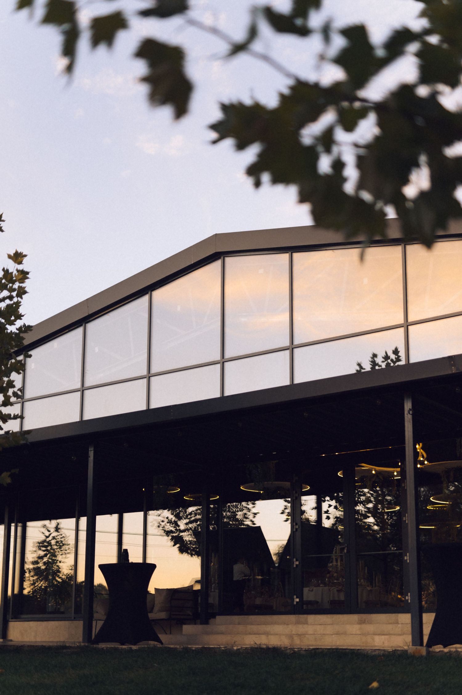 Modern glass pavilion with angular roof silhouetted against sunset sky with tree branches framing the view.