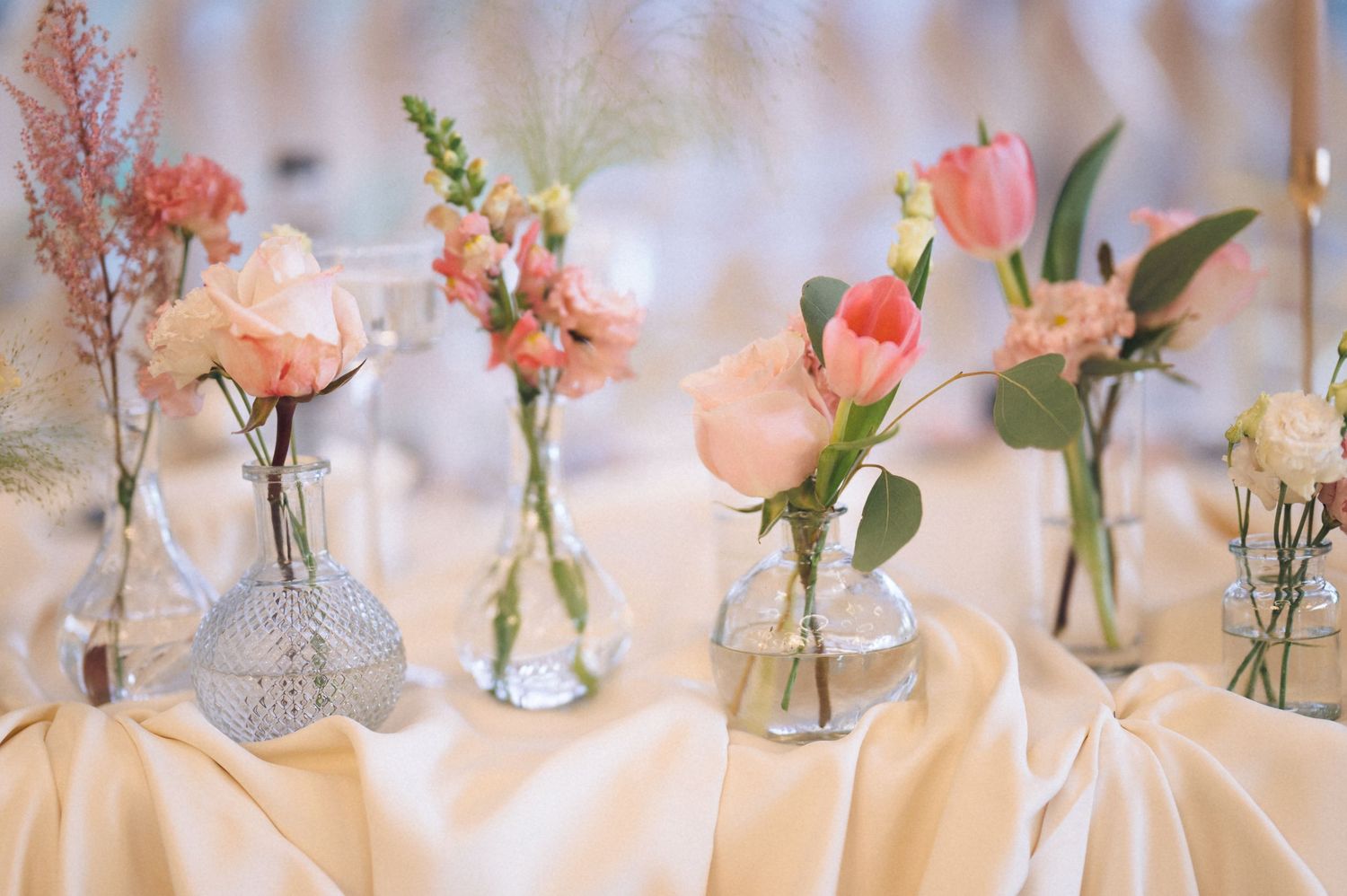 Small glass vases with delicate pink roses and coral flowers arranged in a row on a white tablecloth.