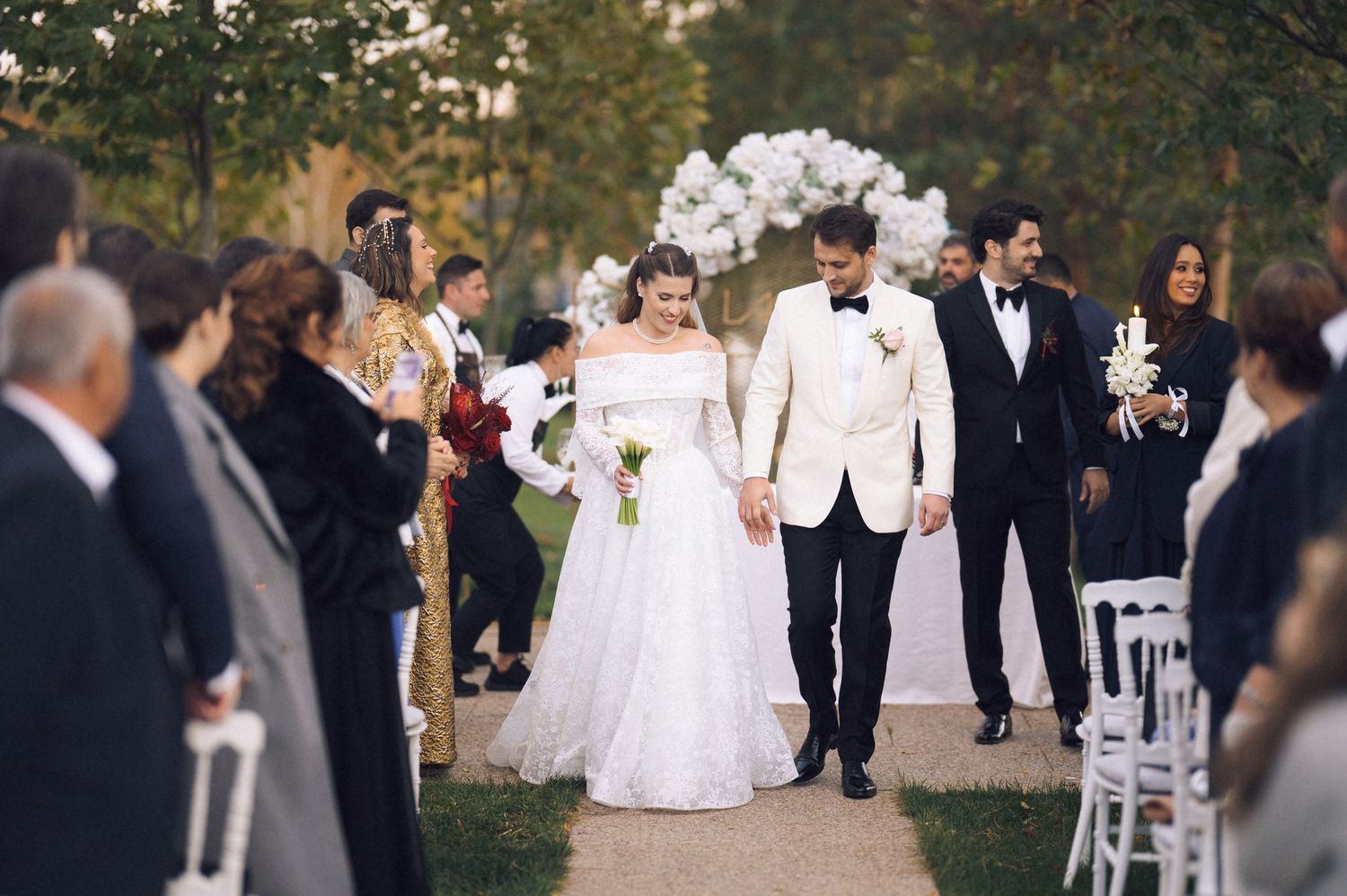 A wedding party walks along a path during an outdoor ceremony in a garden setting.