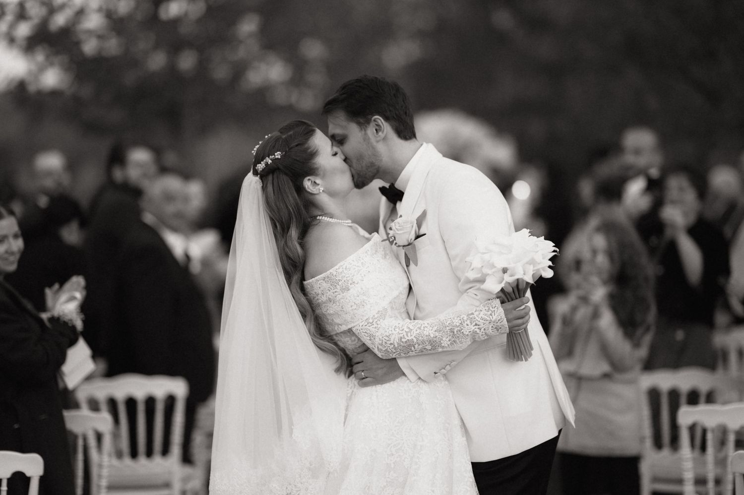 A black and white photograph captures a romantic first kiss between newlyweds at an outdoor wedding ceremony.