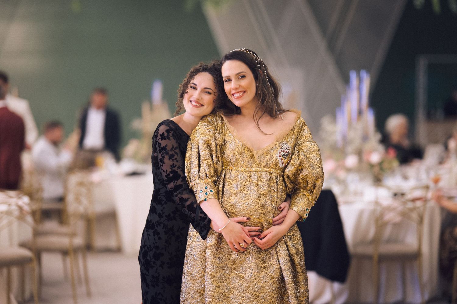 Two guests share a joyful moment at an elegant wedding reception, with one wearing a stunning gold gown.