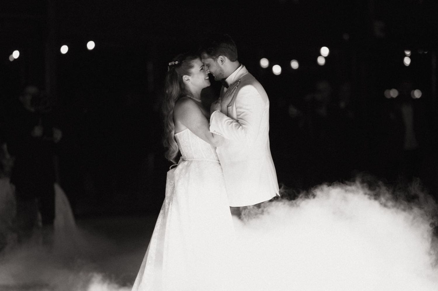 A couple shares their first dance as newlyweds on a misty dance floor with twinkling lights in the background.