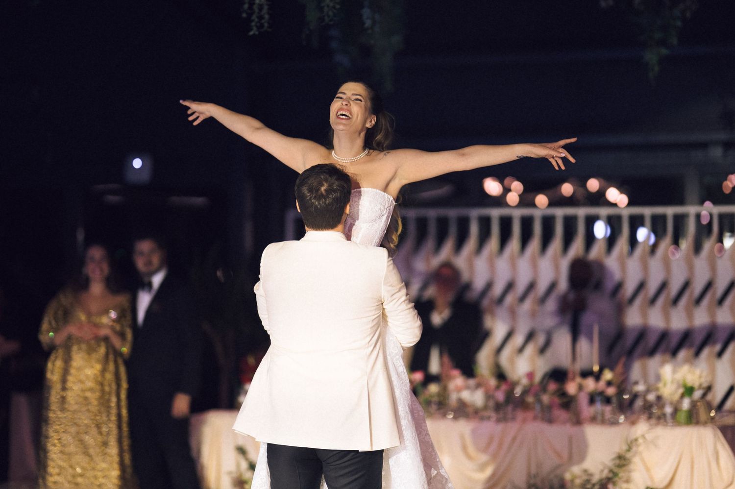 A couple dances gracefully at a formal evening event with decorative railings and lights in the background.