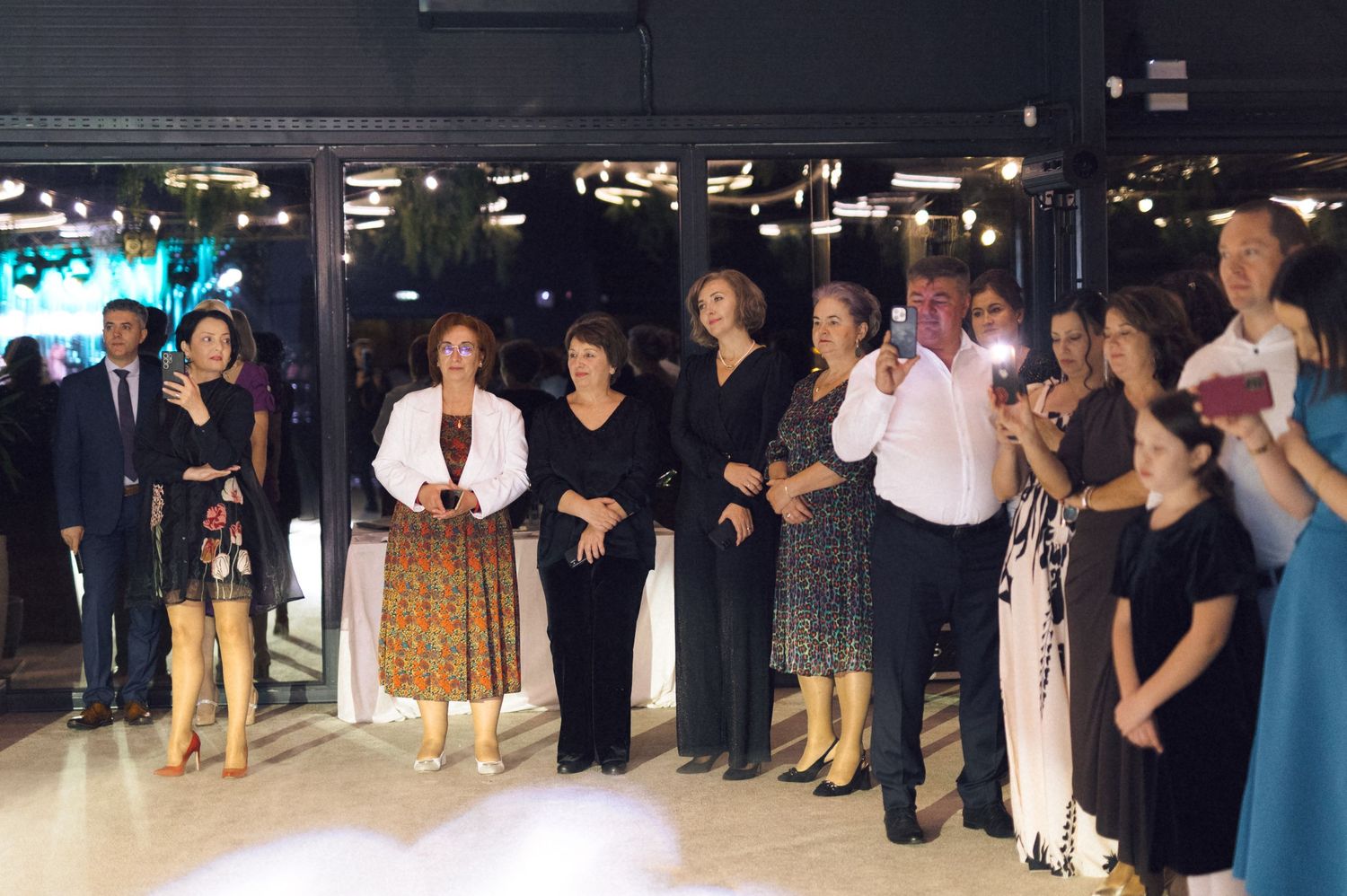 A group of people in formal attire stand together in a line at an indoor event venue with dark lighting.