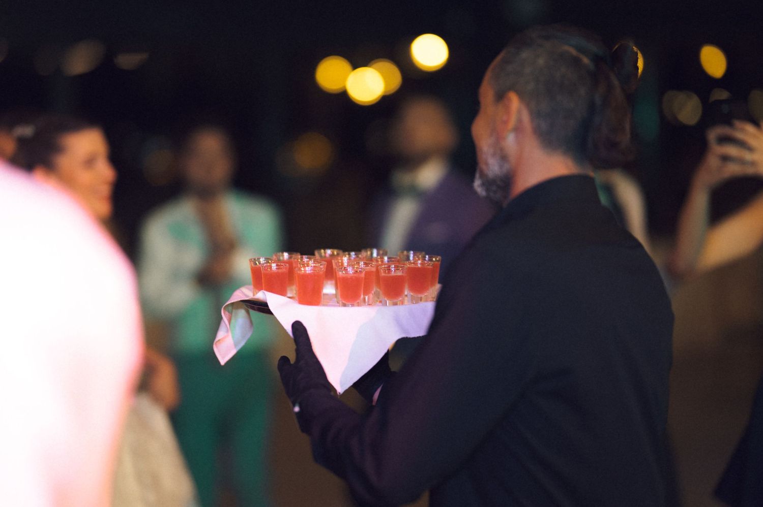 Server carrying a tray of red cocktails in martini glasses at an evening event with bokeh lights in background.