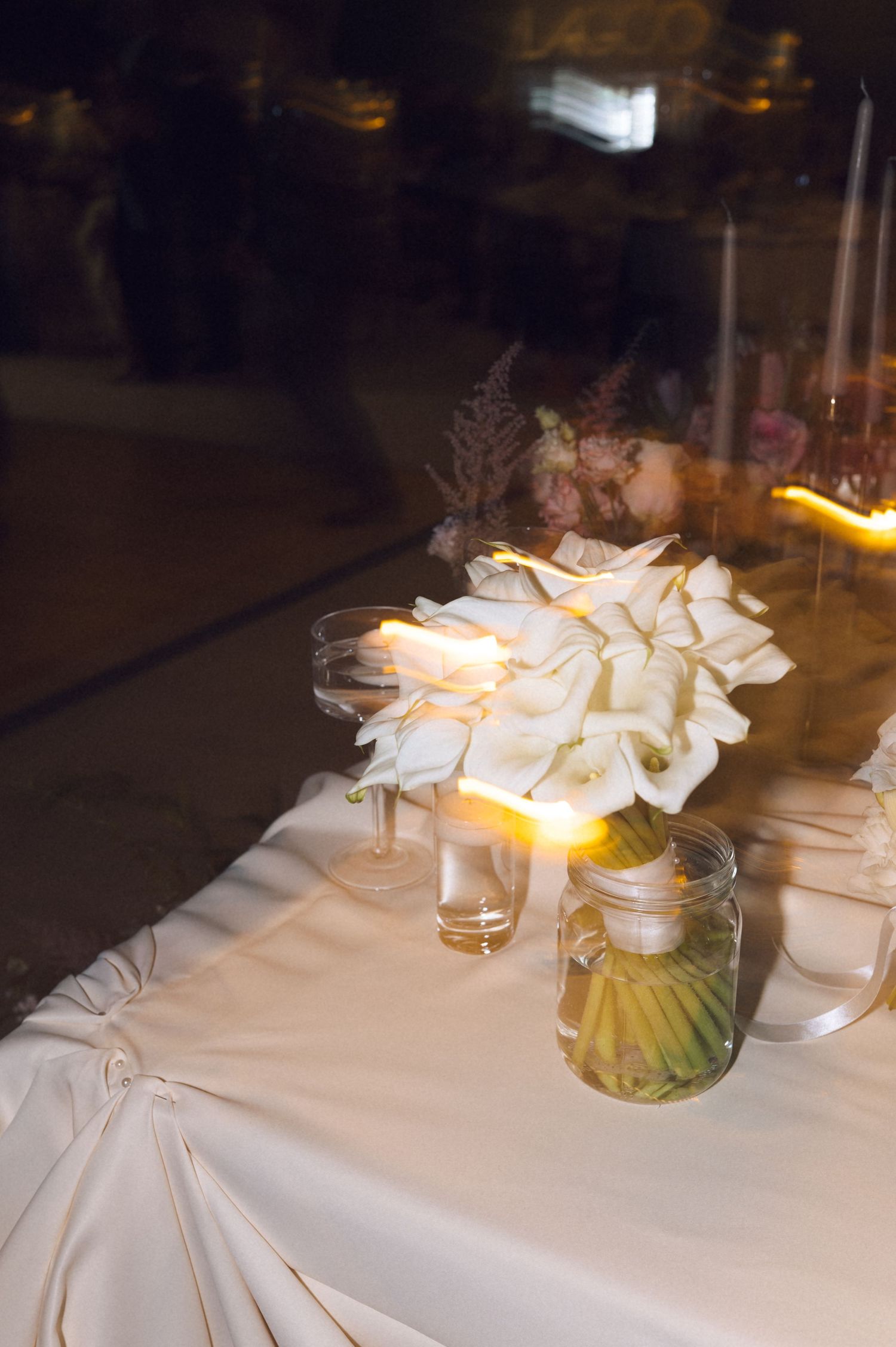 Elegant table setting with white linens, glowing candles and floral arrangements in glass vases at an evening event.