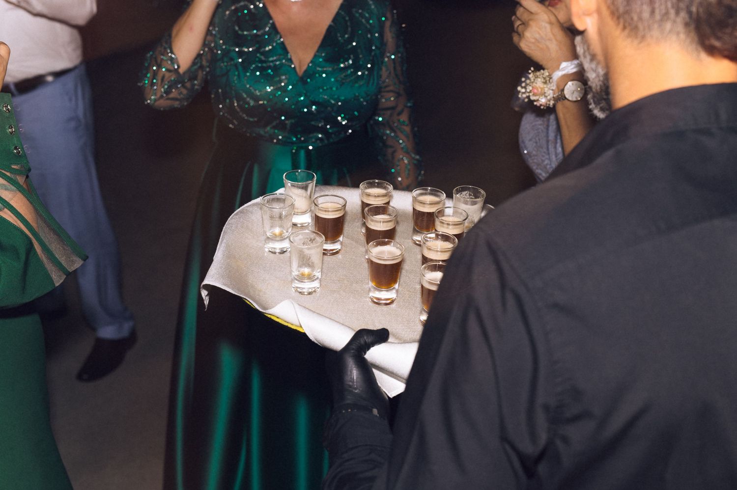 Server holding tray of layered coffee drinks in shot glasses at formal event with emerald green dress visible.