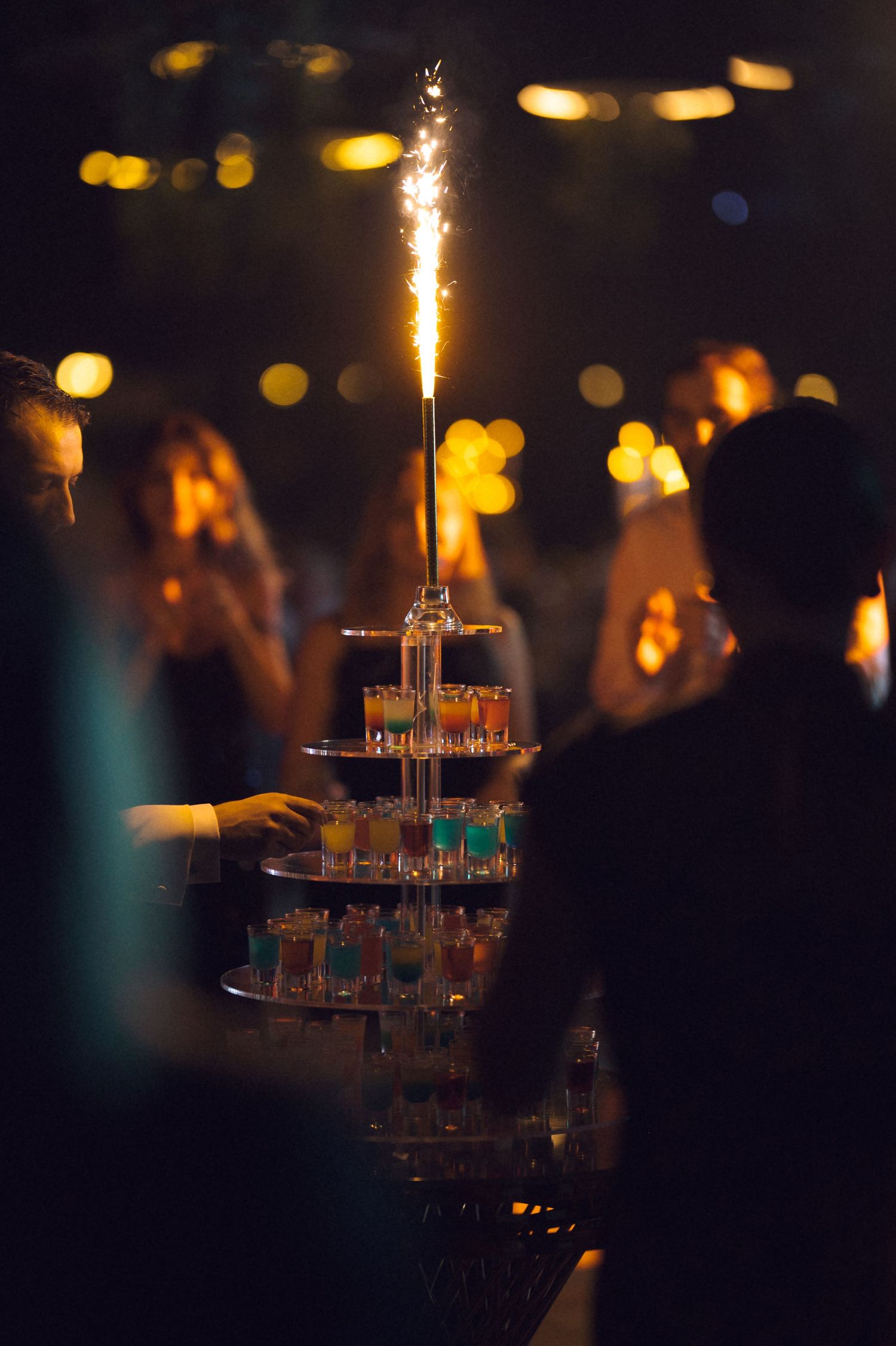 A sparkler candle illuminates a tiered display of champagne glasses at a dimly lit celebration event.
