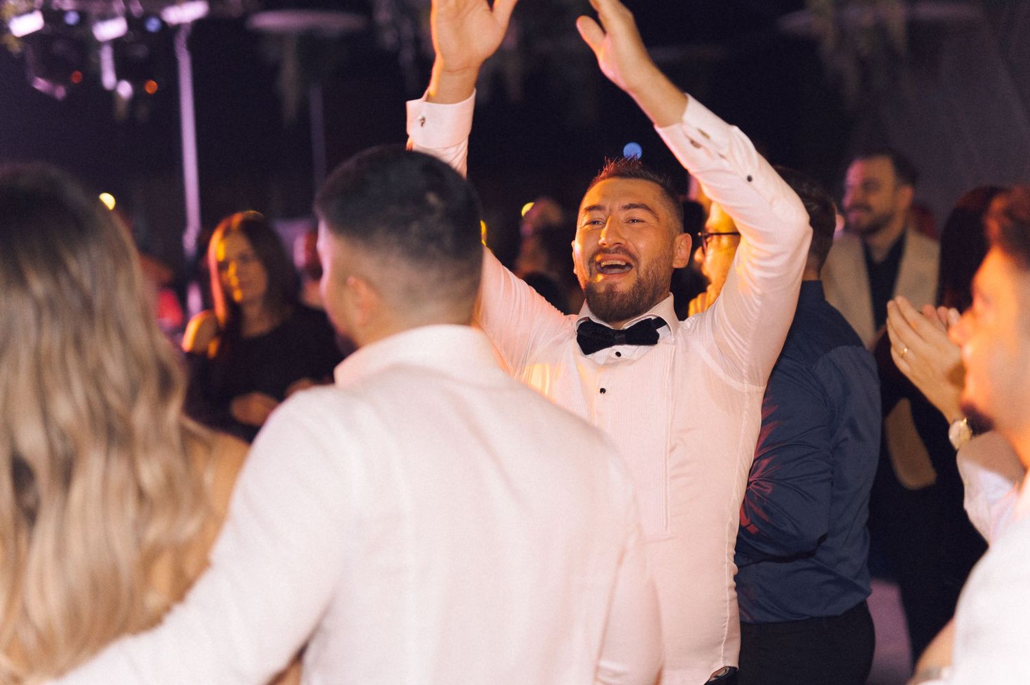 People dance and celebrate at a wedding reception in formal attire with raised arms and joyful expressions.