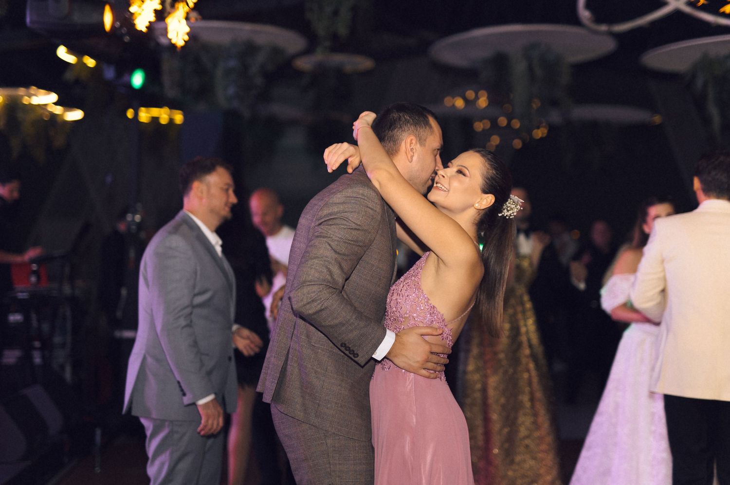A couple shares a romantic dance at an evening reception, with the woman wearing a pink dress and man in a gray suit.