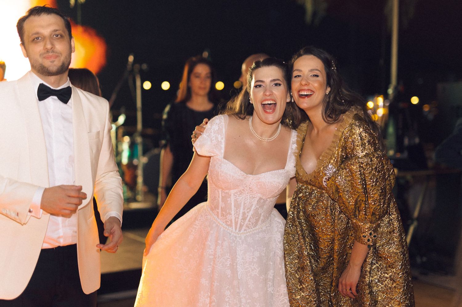Wedding guests dance and celebrate at an evening reception in formal attire with glowing lights in the background.