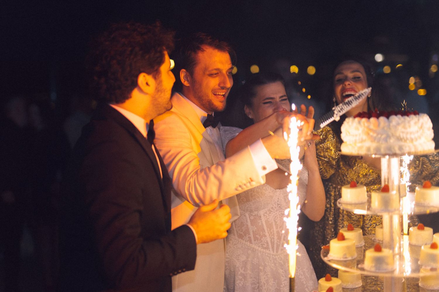 Guests gather around a glowing wedding cake with sparklers at an evening reception celebration.