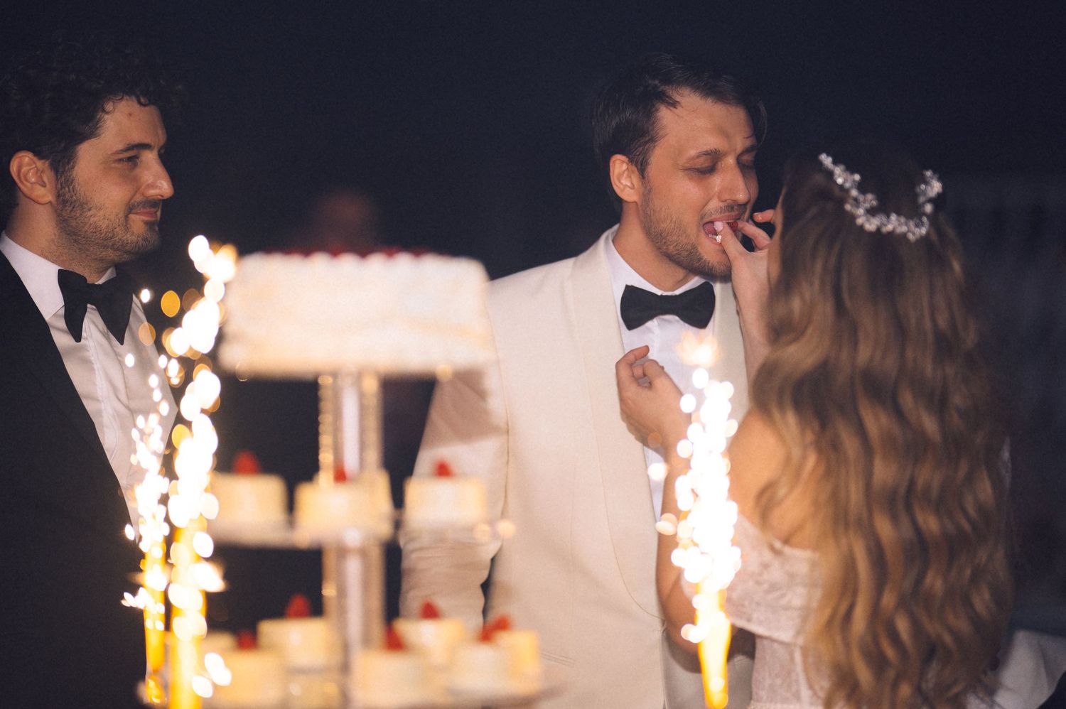 A romantic wedding reception scene with sparklers and a wedding cake in the background during an evening celebration.