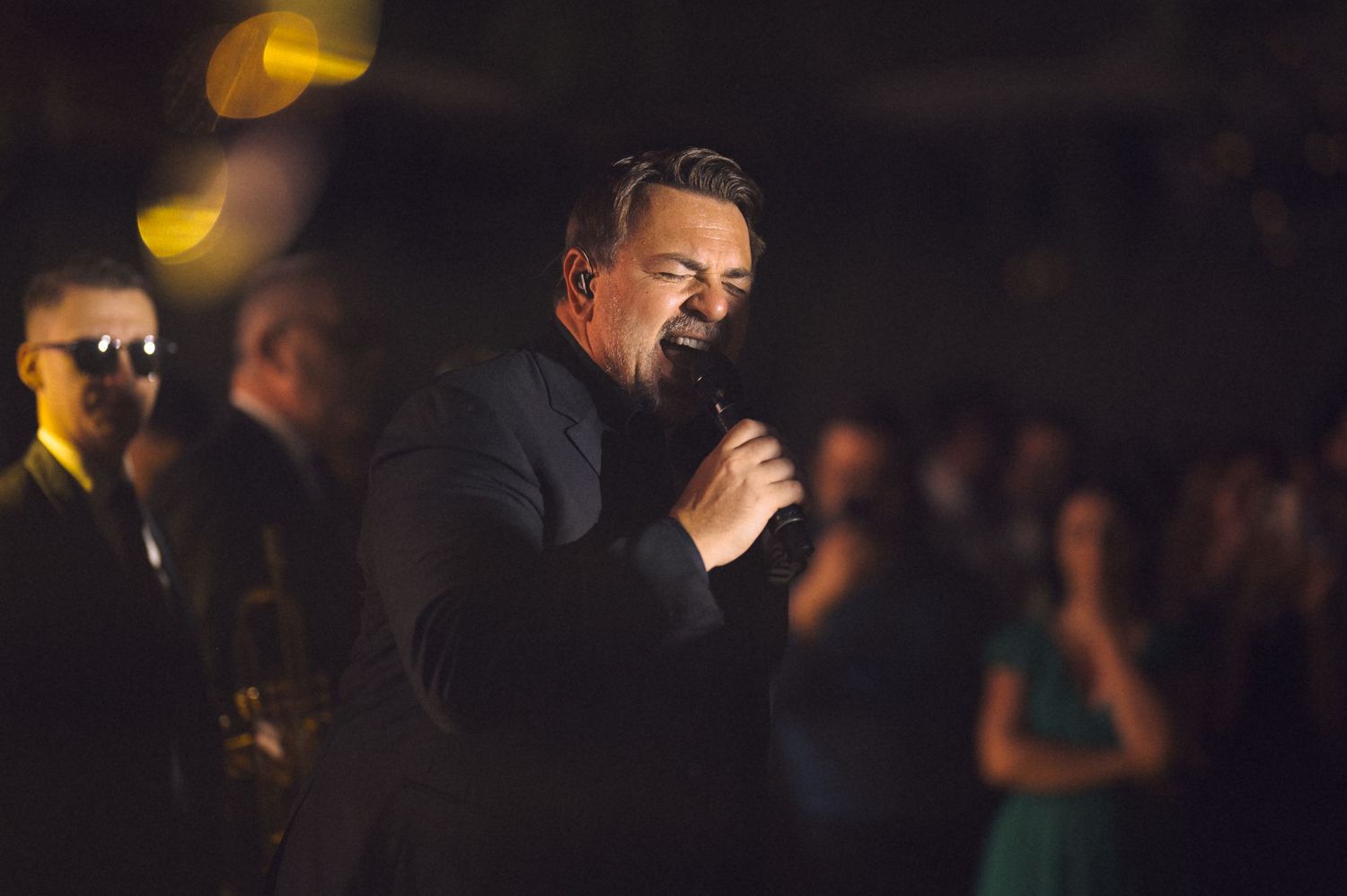 A performer sings into a microphone on a dimly lit stage with warm lighting and audience members visible in the background.