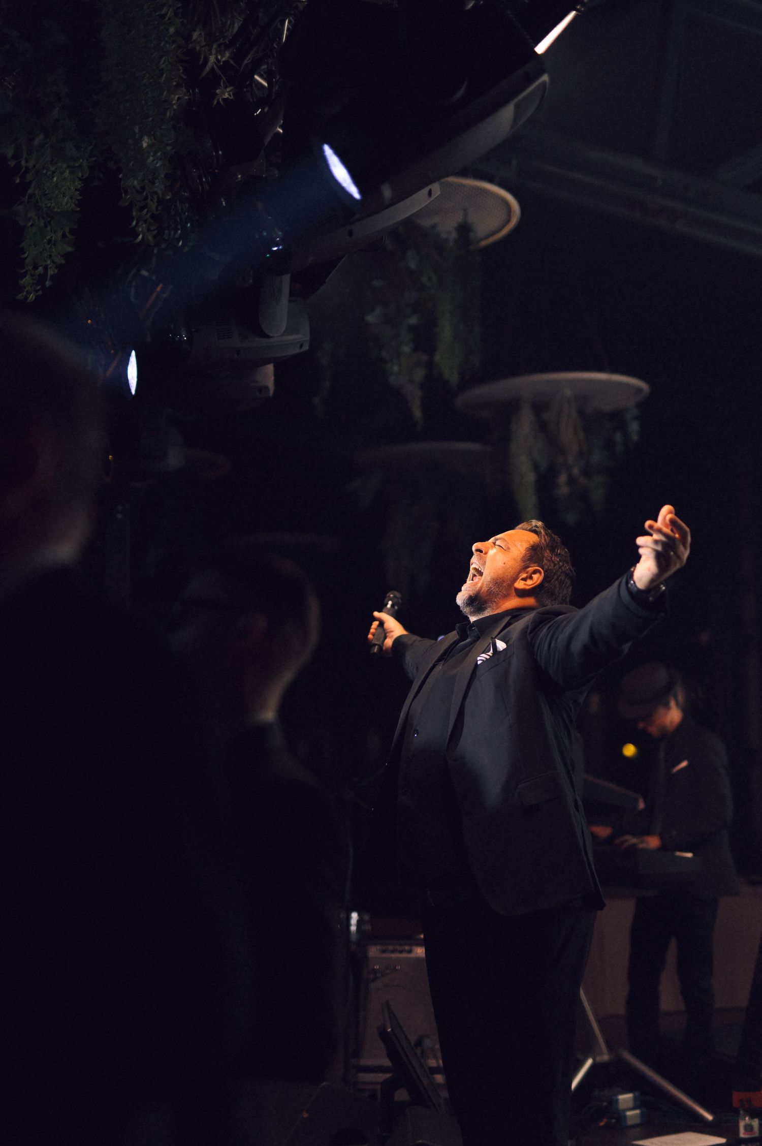 A performer in black attire sings passionately on stage with dramatic lighting and raised arms during a theatrical show.