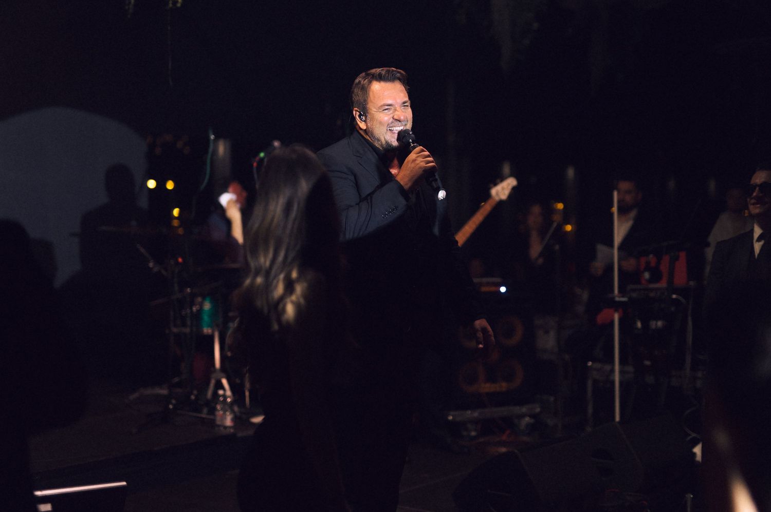 A performer sings passionately on a dark stage with musicians and instruments visible in the background.