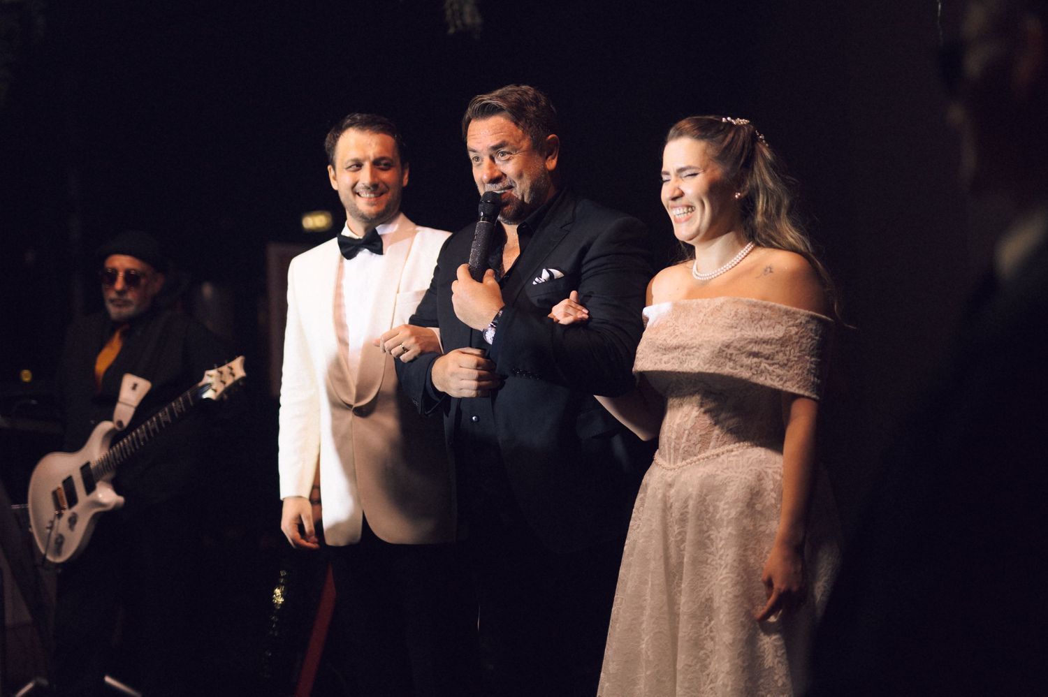 Three performers on stage during an elegant evening performance with a guitarist visible in background.