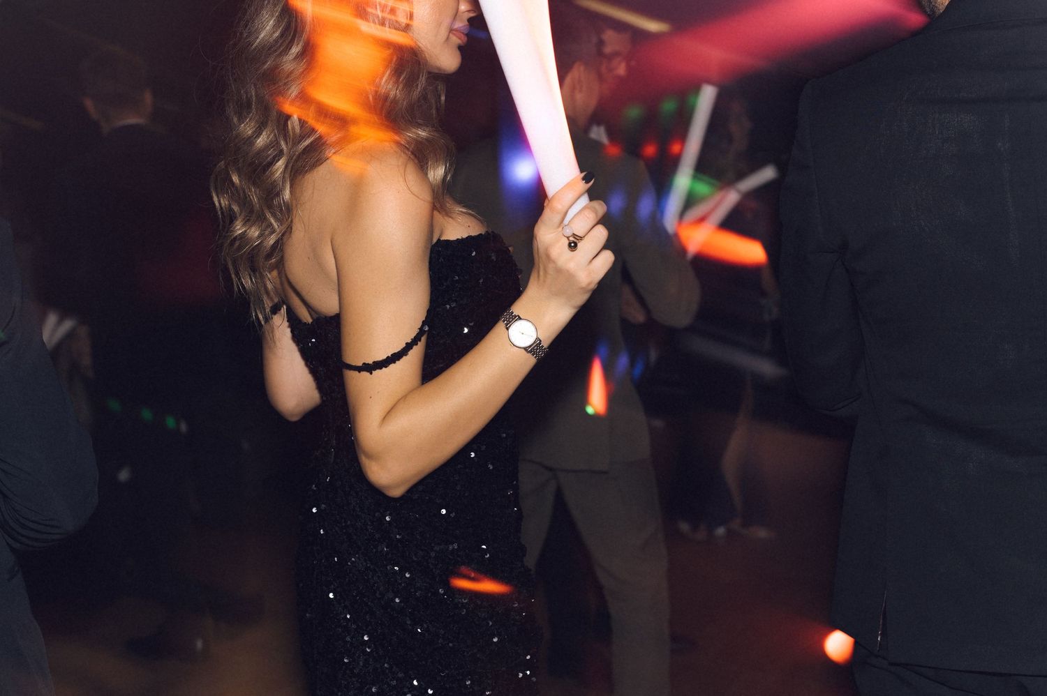 A person in a sparkly black dress holds a glowing light stick while dancing at a party.