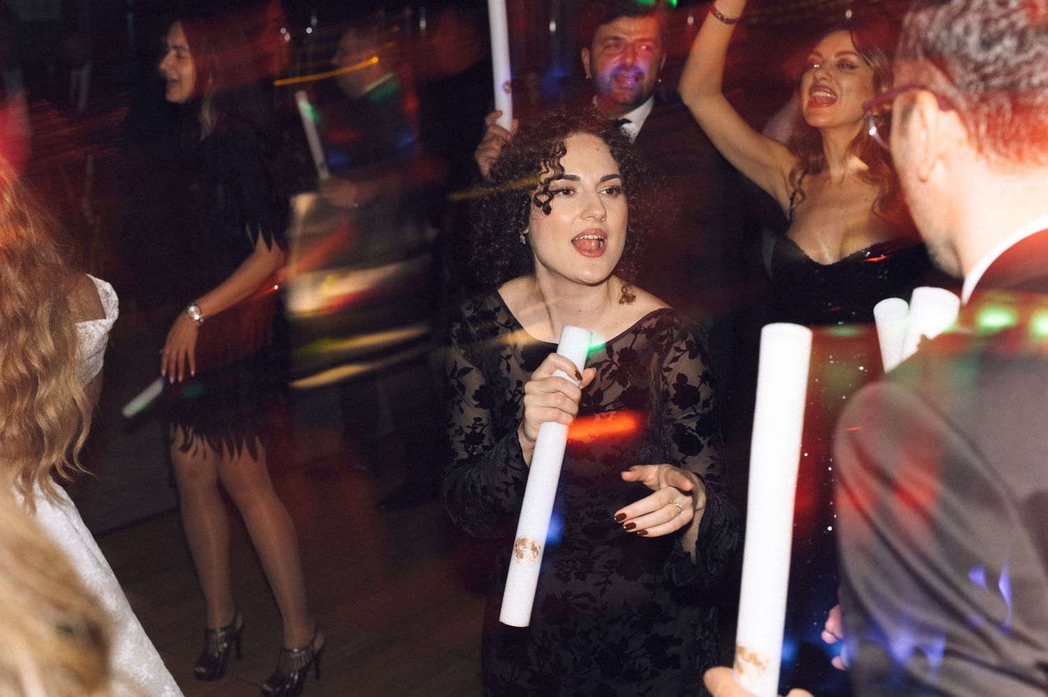 Party scene with people dancing and holding glowsticks at a nightclub with colorful lighting effects.