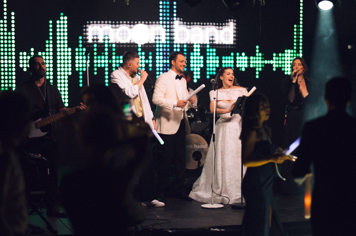 Performers on stage at a nightclub with green digital equalizer display backdrop and spotlights illuminating the scene.
