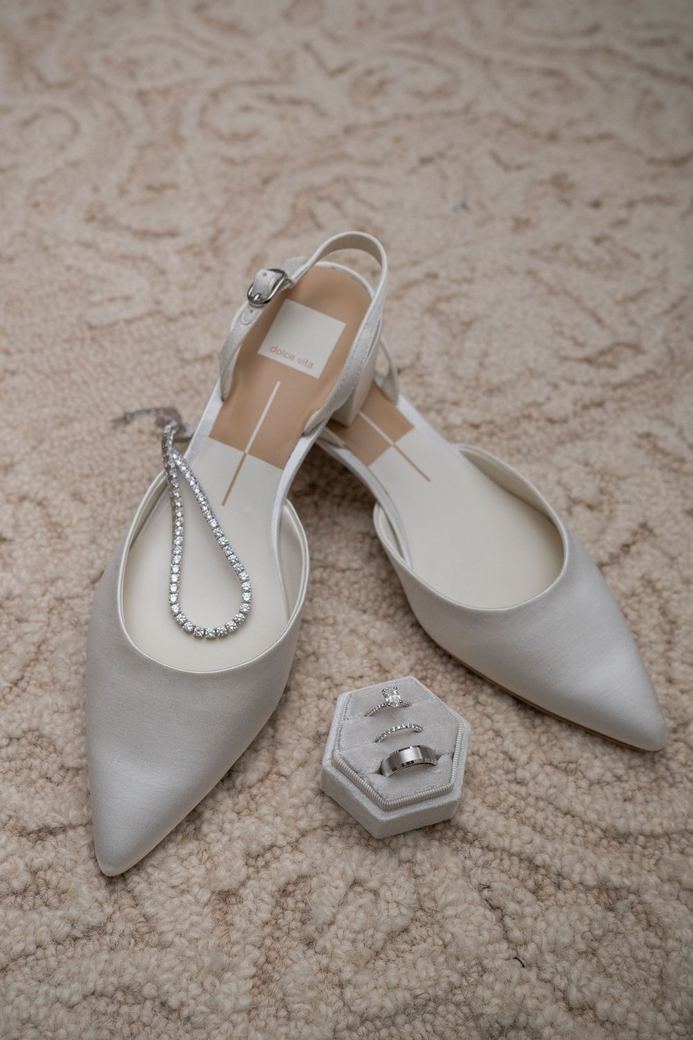 Elegant silver slingback wedding shoes with crystal details displayed on a beige carpet alongside a hexagonal ring box.