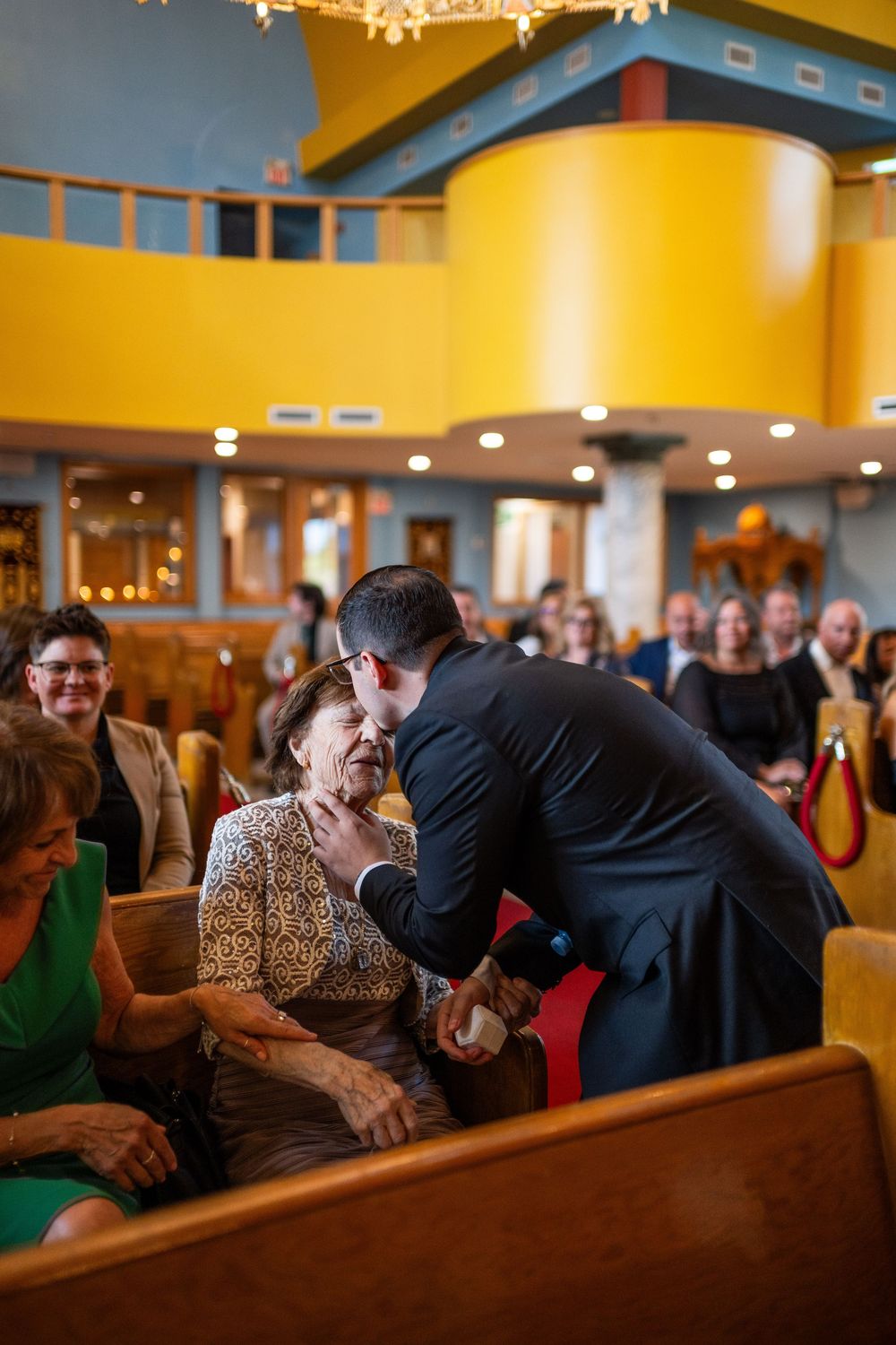 Wedding guests share joyful moments in wooden church pews with warm yellow lighting and intimate atmosphere.