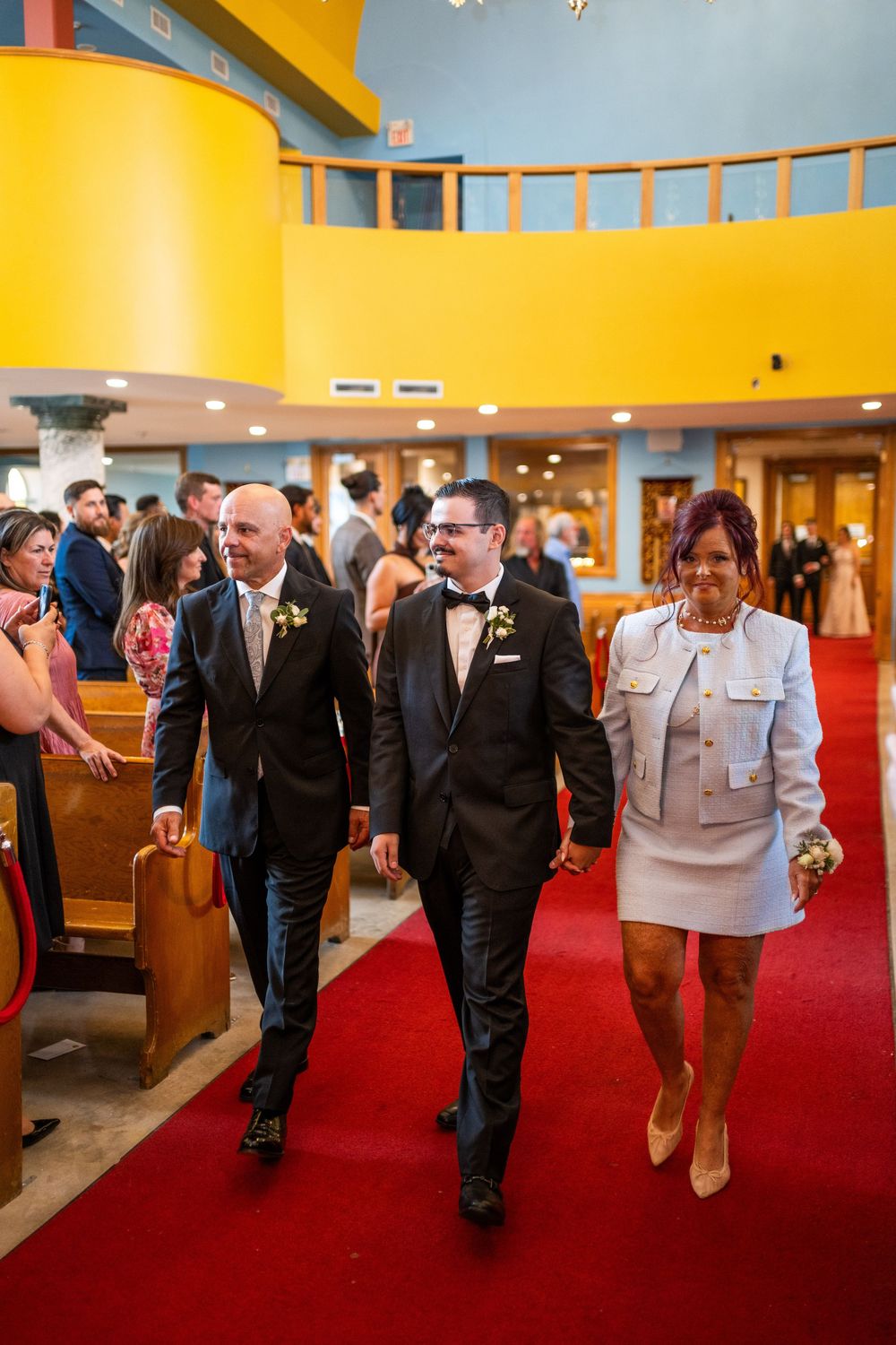 Wedding party members walk down a red carpeted aisle in a modern church with vibrant yellow and blue walls.