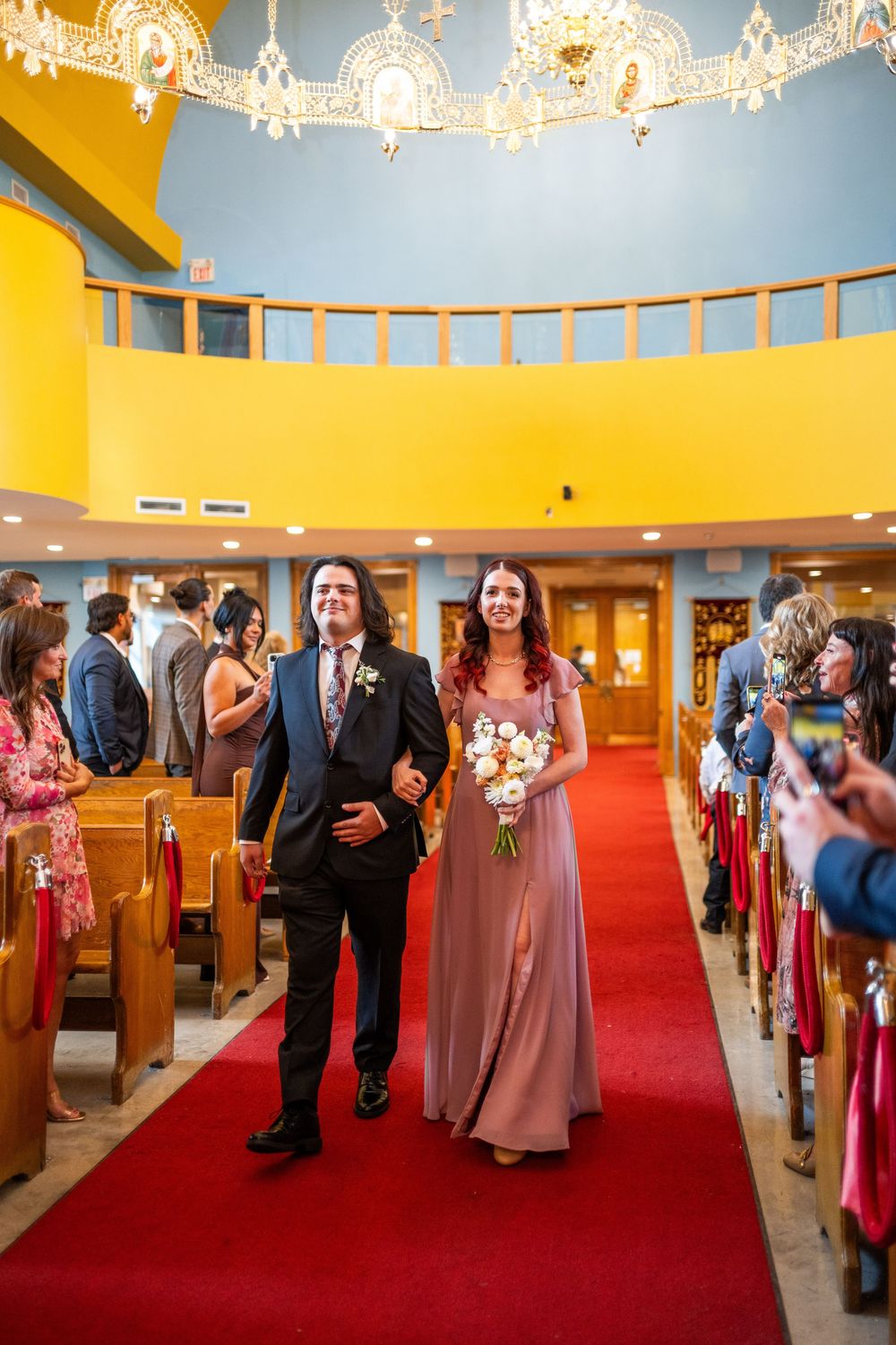 Wedding ceremony aisle view with red carpet and guests gathered in a church illuminated by elegant chandeliers.