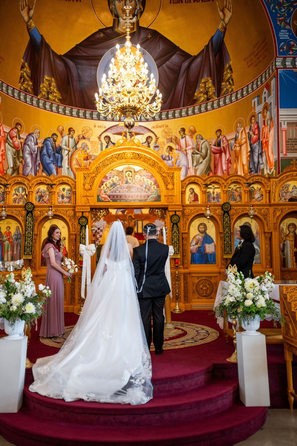 Orthodox wedding ceremony taking place in an ornate church with golden walls and religious icons under a crystal chandelier.