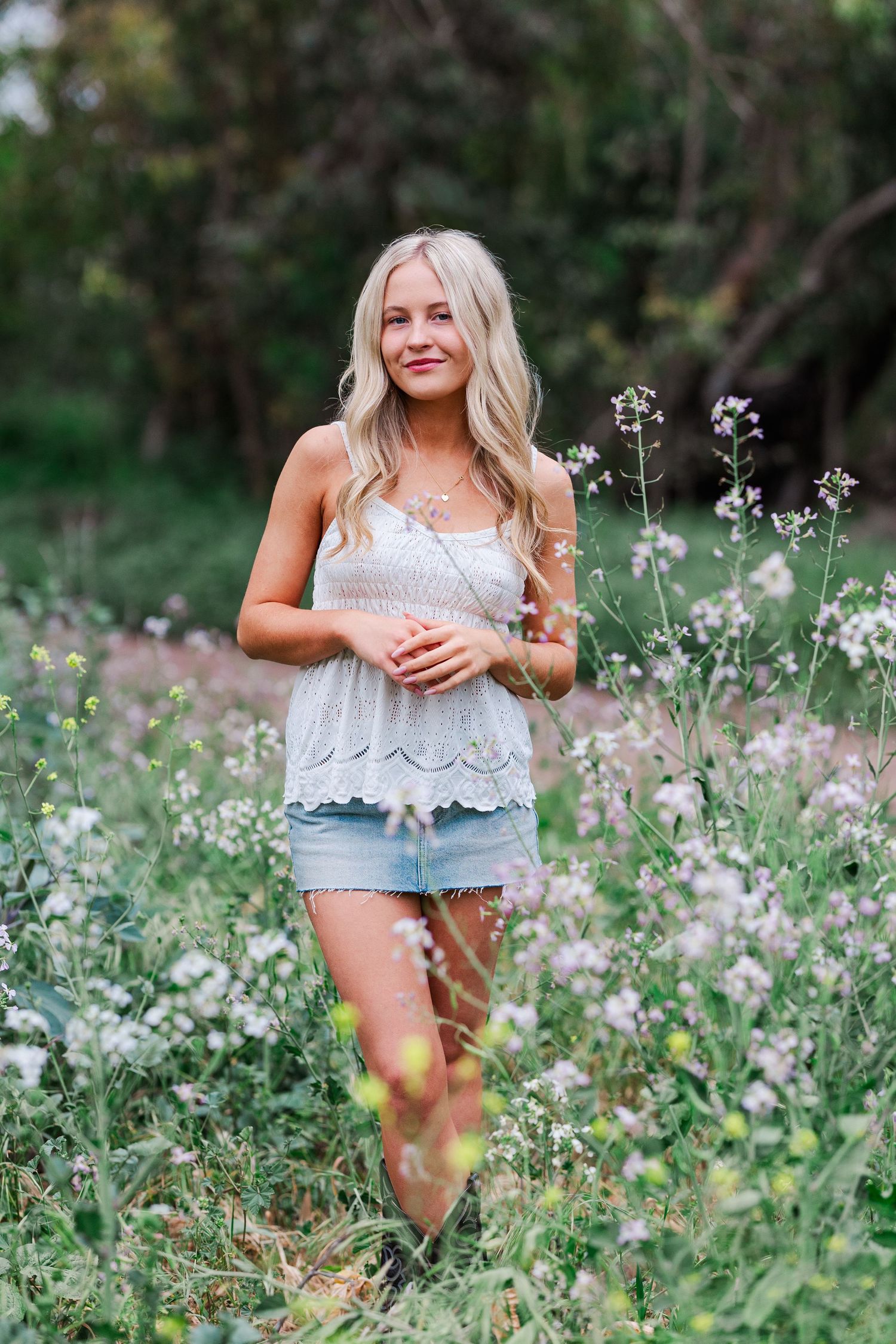 A person in a white lace top and denim shorts poses playfully in a meadow of delicate white wildflowers on a sunny day.