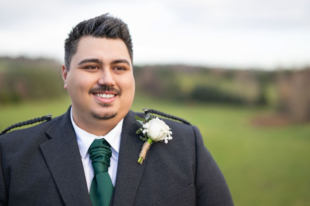 Groomsmen in dark suits with green ties and white boutonnieres pose outdoors on a grassy hillside.