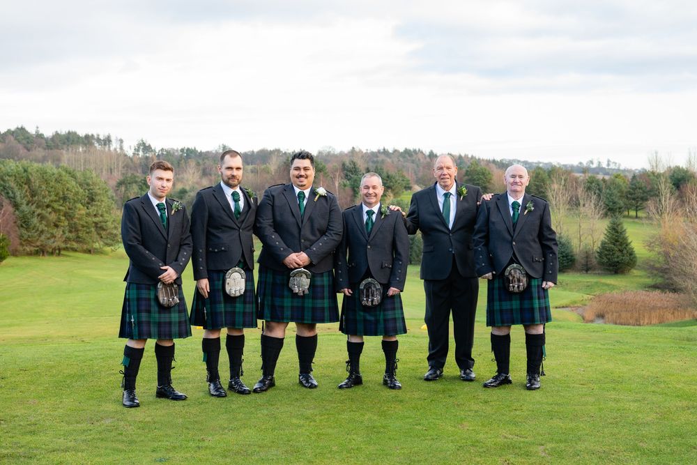 Wedding party in formal attire lined up sitting on grass with scenic countryside views in background.