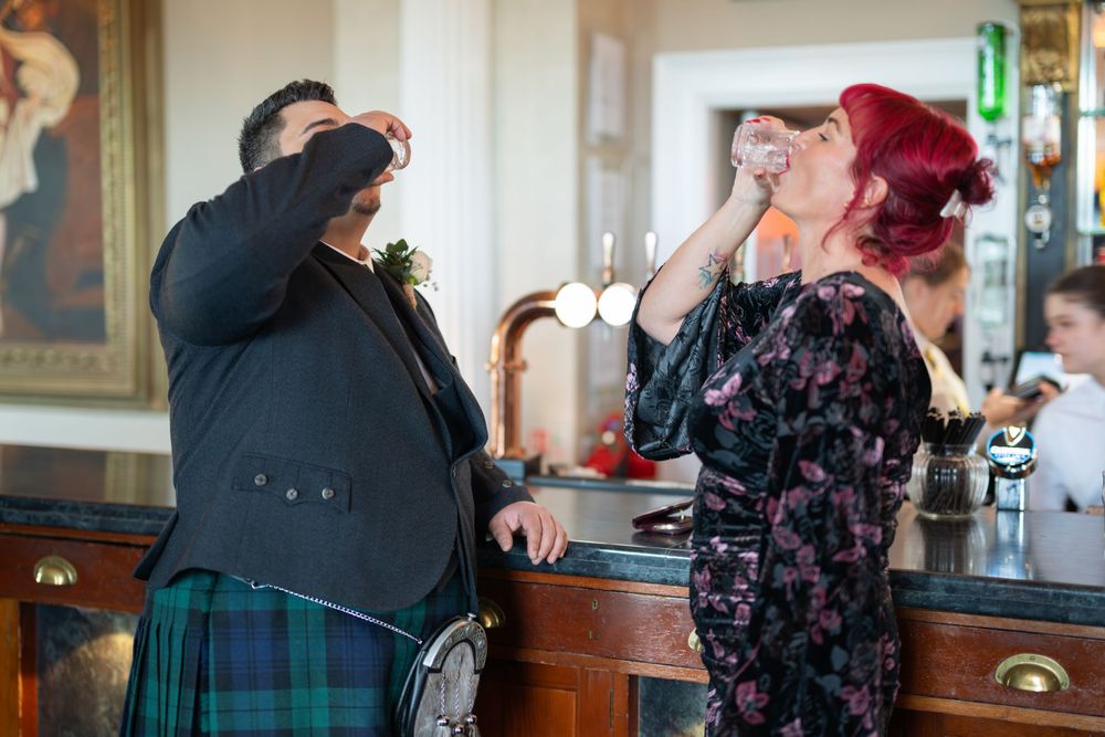 Two people share a playful moment at a bar wearing traditional Scottish attire and formal clothing.