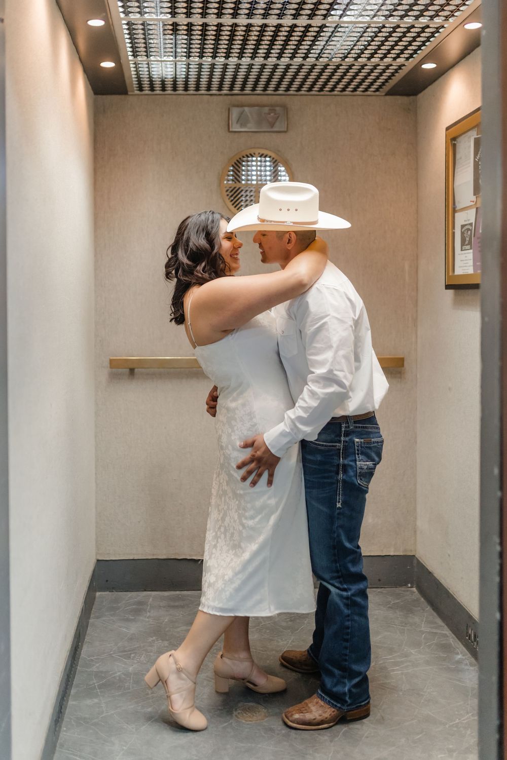 A couple sharing a romantic moment in a hallway, with one wearing a white dress and the other in cowboy attire.