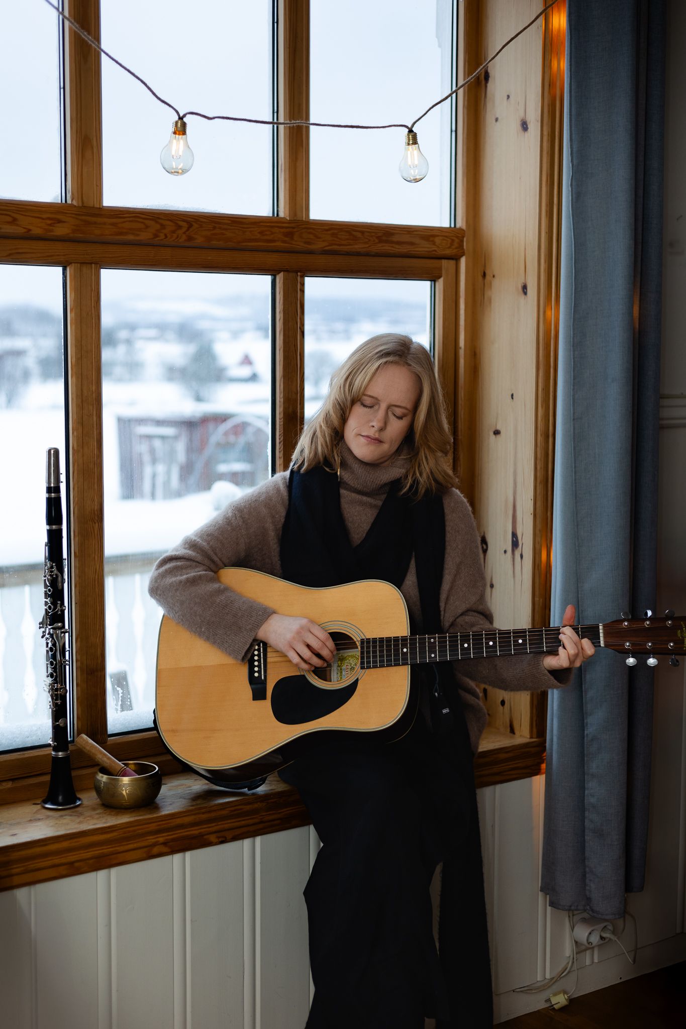 Musician plays acoustic guitar by window with string lights above and snowy winter scene visible outside.