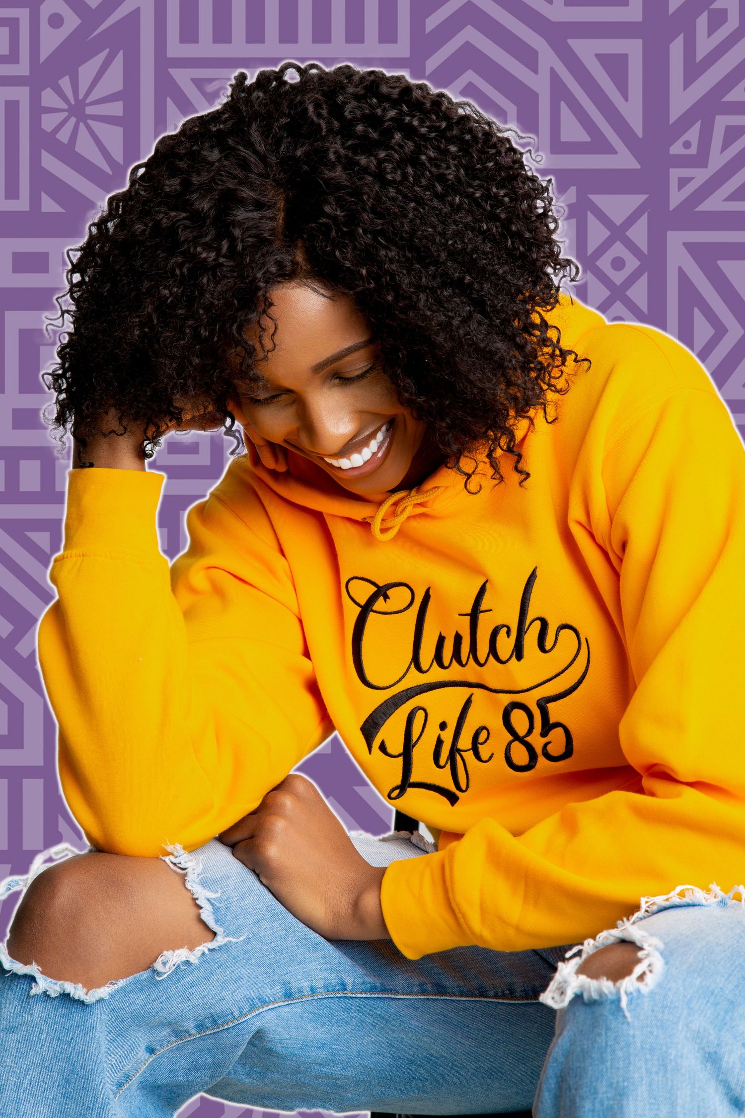 Woman smiling in yellow Clutch Life 85 hoodie and ripped jeans on purple patterned background.