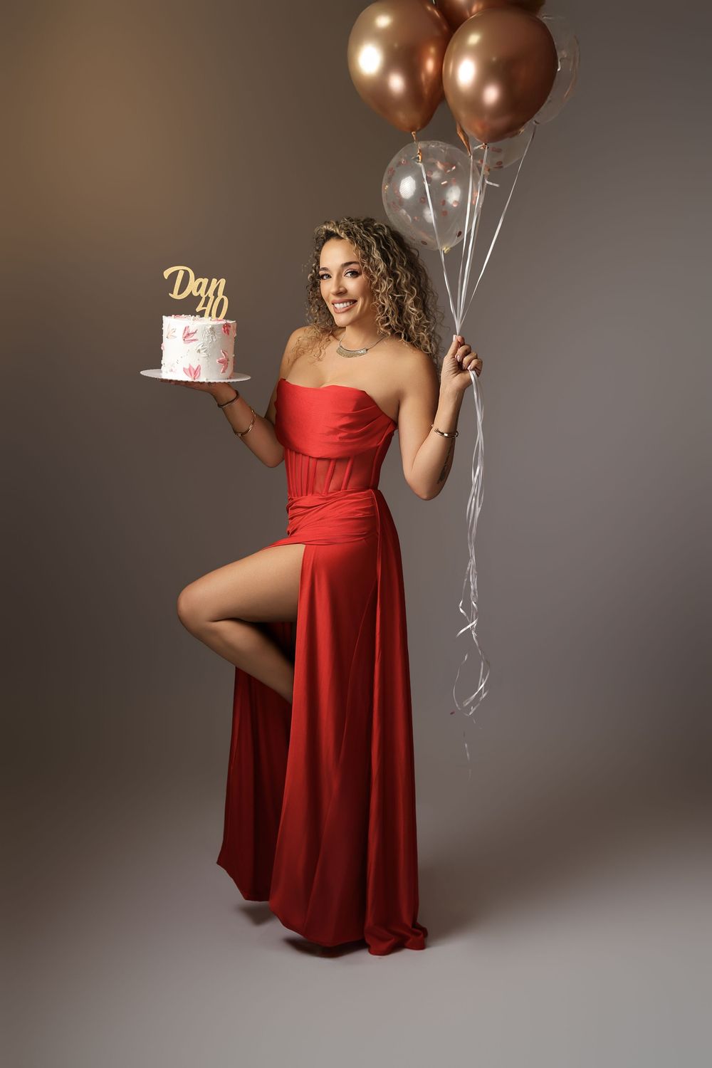 Glamorous photoshoot featuring a red strapless dress and rose gold balloons against a grey backdrop.