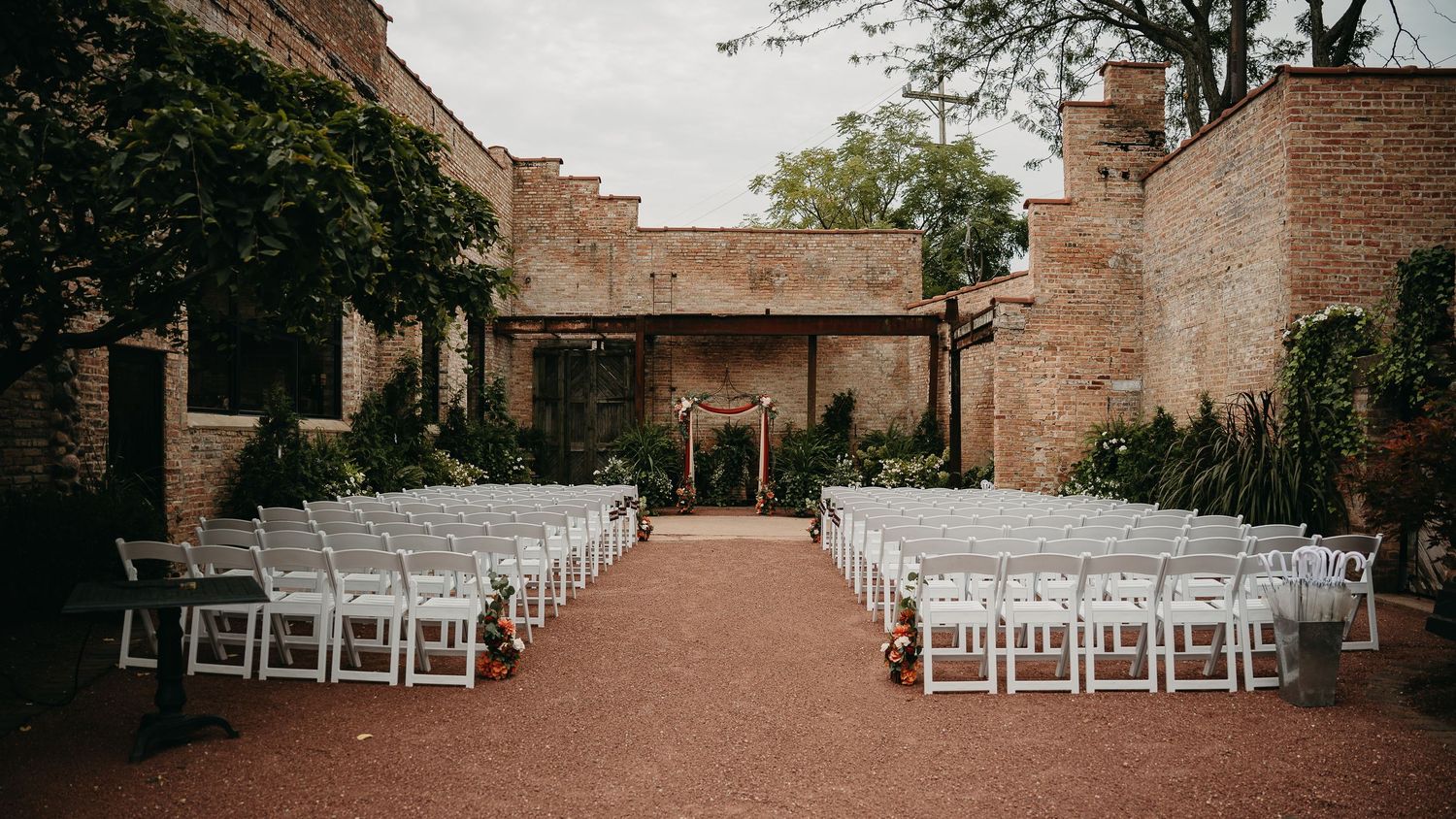A panoramic view of a rustic brick courtyard set up for an outdoor wedding ceremony with white chairs and greenery.